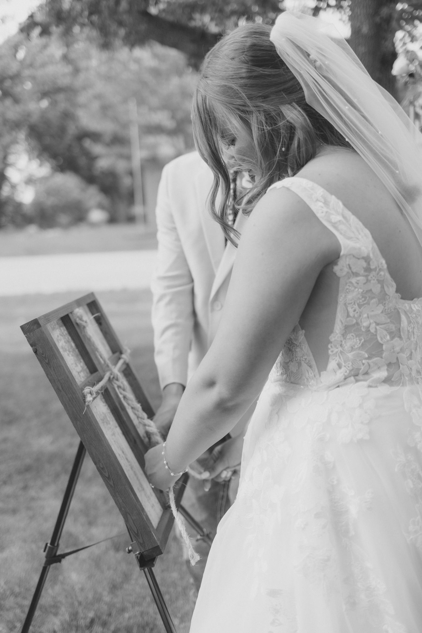 Bride in lace wedding gown signs guest book on wooden easel at outdoor wedding ceremony.