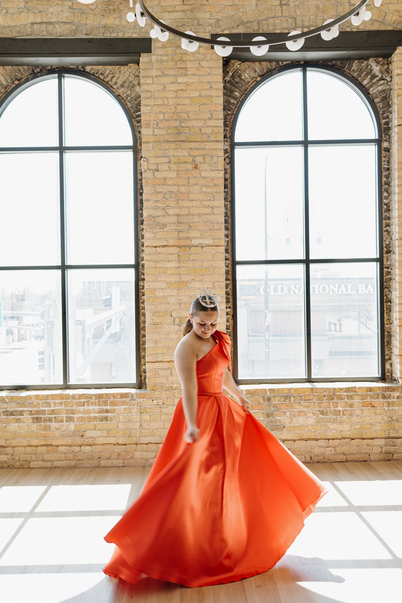Woman twirling in flowing red one-shoulder ball gown in sunlit brick loft.