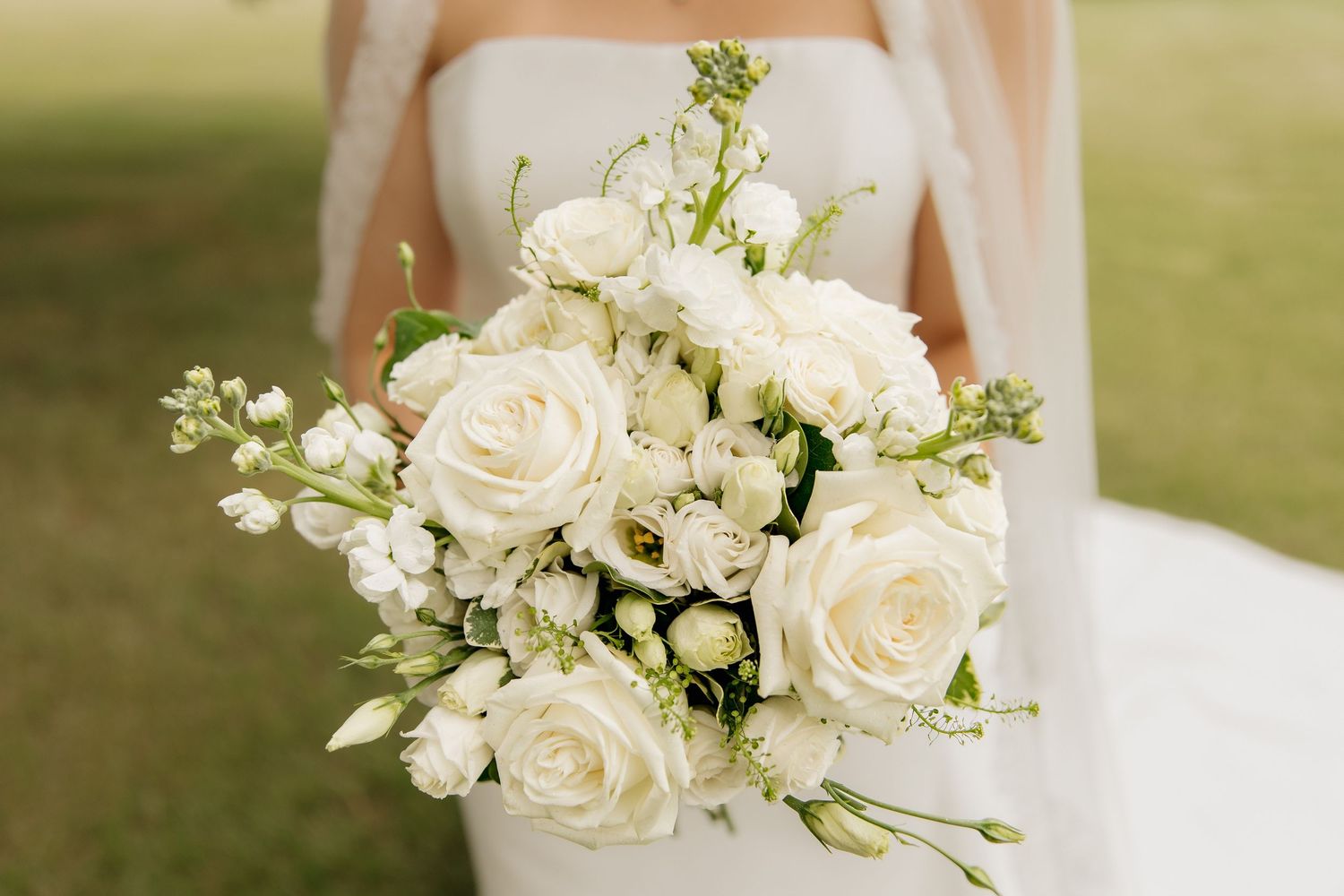 Bride holding a lush white wedding bouquet of roses and lisianthus against a white gown outdoors.