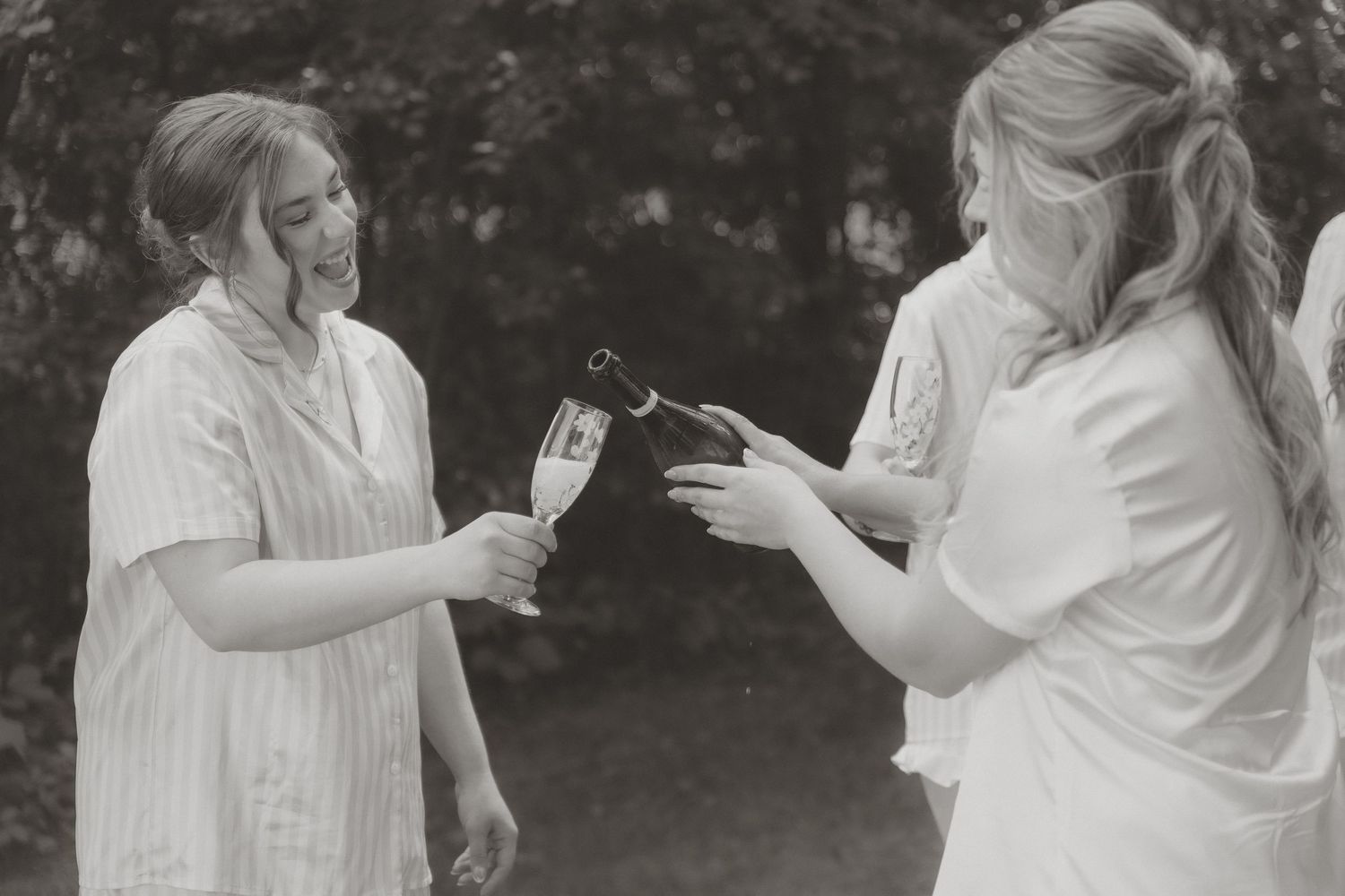 Black and white photo of two women in white dresses pouring champagne into a glass outdoors.