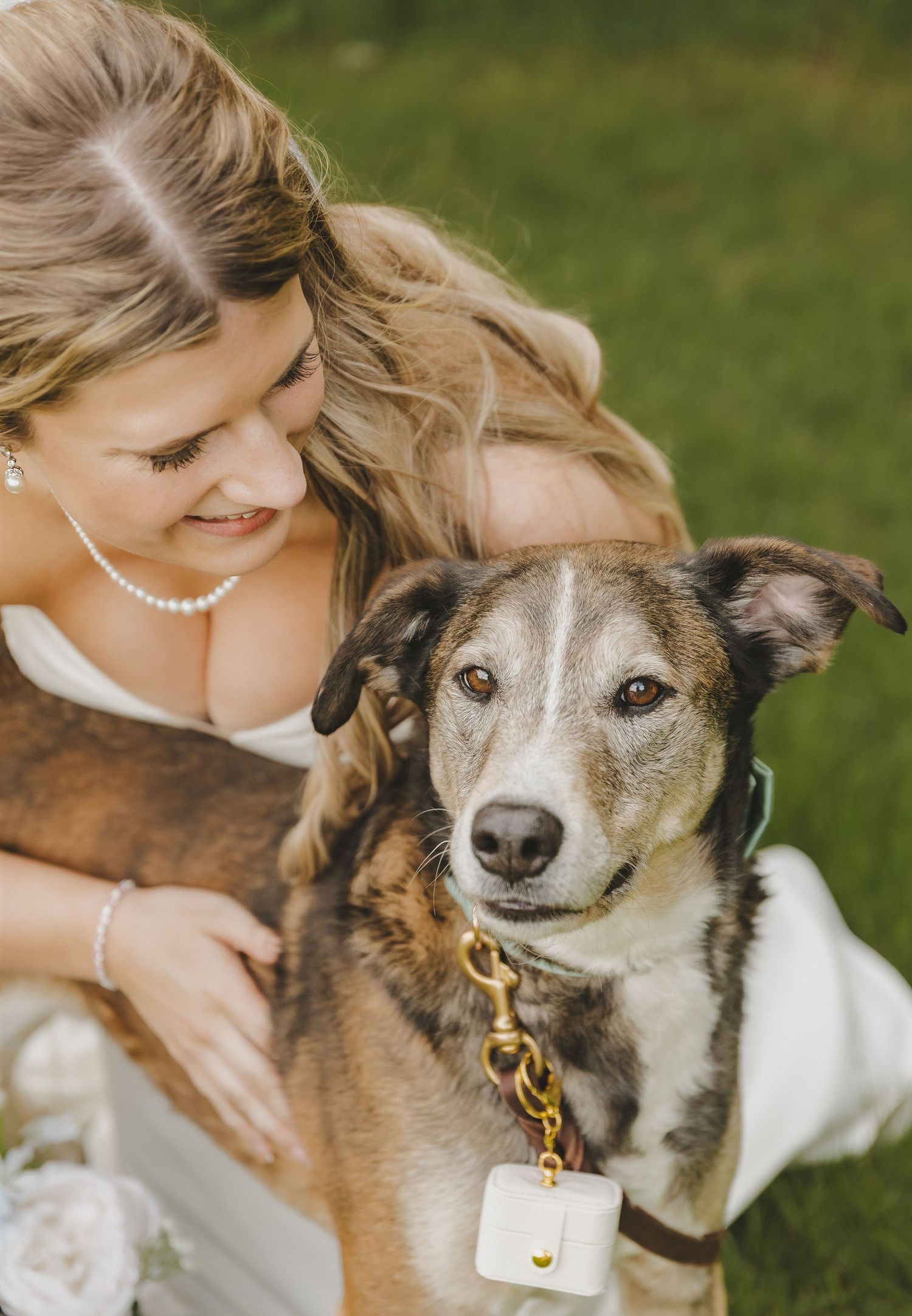 Smiling bride in white gown cuddling a brown and gray dog wearing a gold chain collar outdoors.