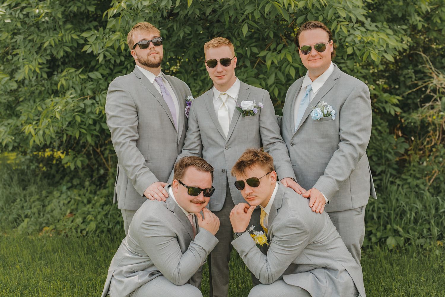 Five groomsmen in matching gray suits and sunglasses posing outdoors in a fun wedding party photo.