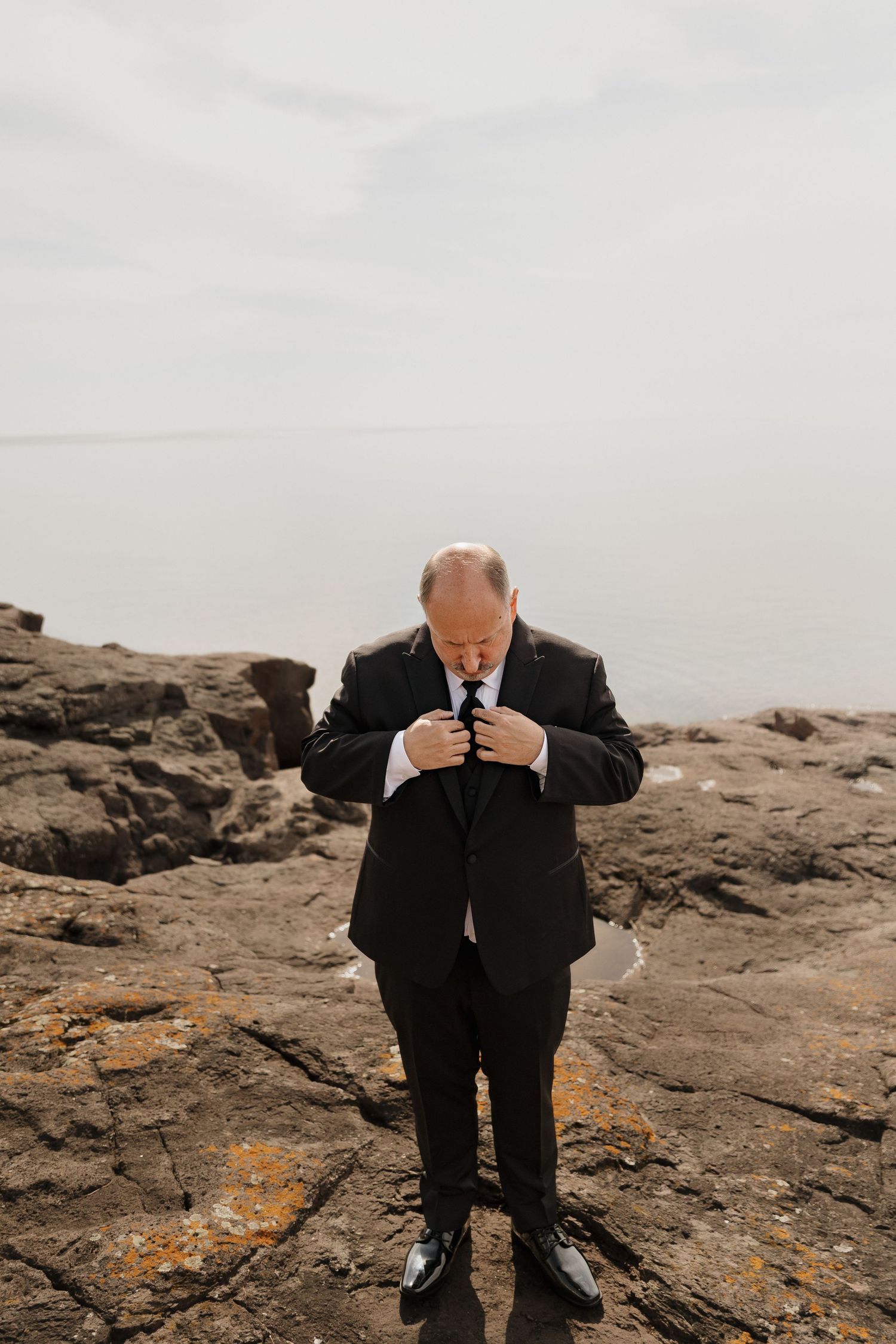 Groom in black suit adjusting his tie on rocky shoreline with overcast sky backdrop.