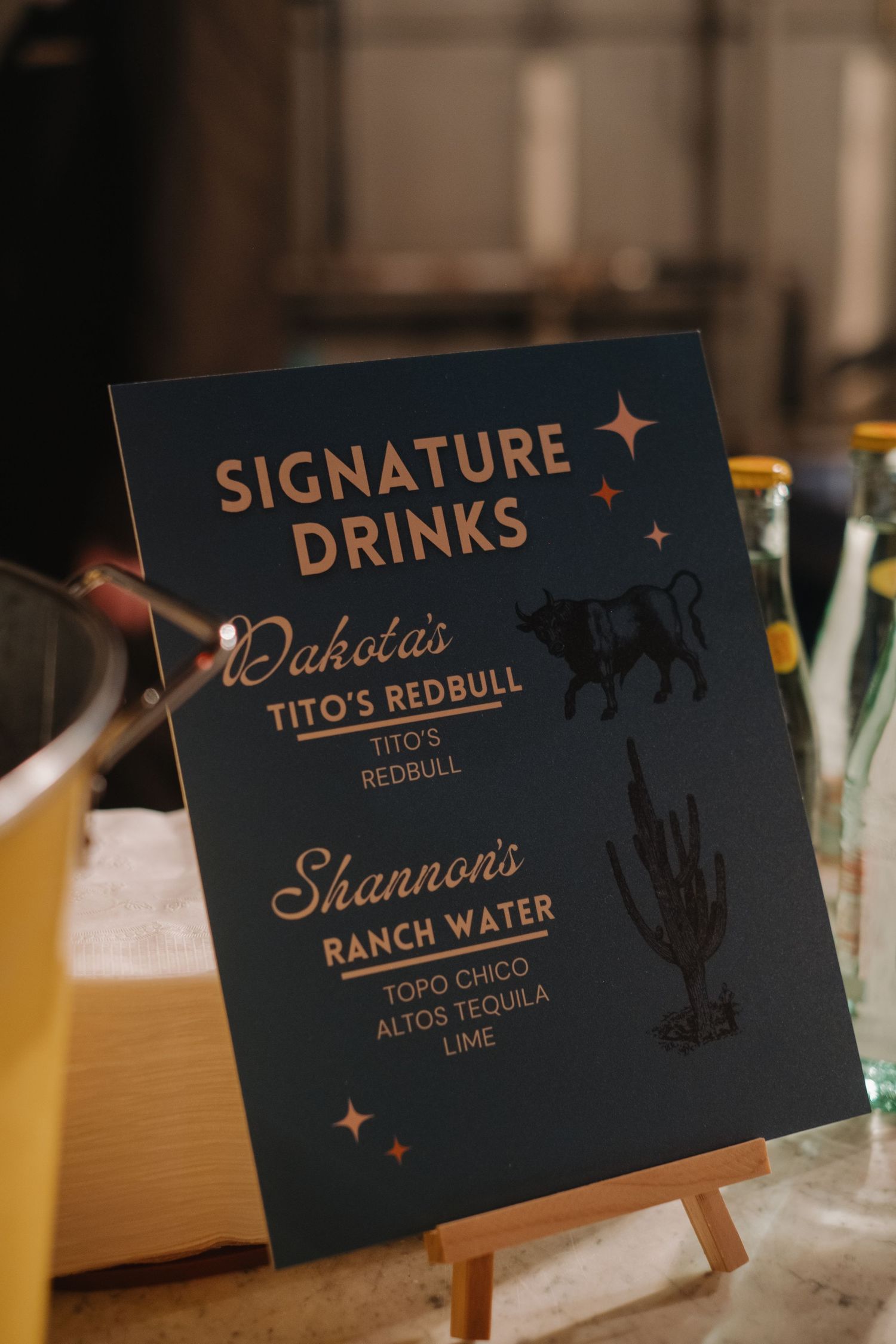 Dark blue wedding signage displaying signature drink menu items with gold lettering.