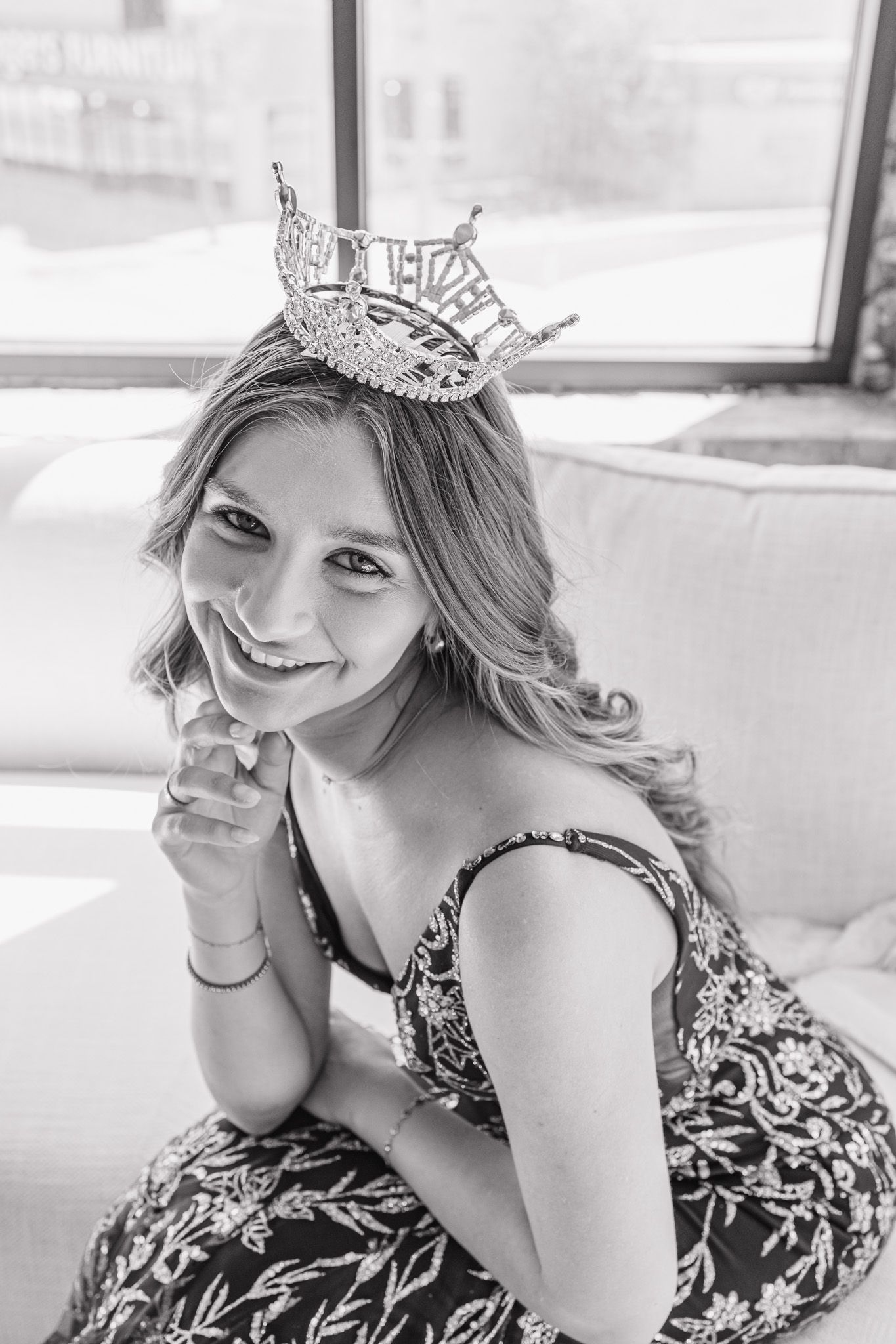Smiling young woman wearing a pageant crown poses gracefully in a floral dress, black and white portrait photo.