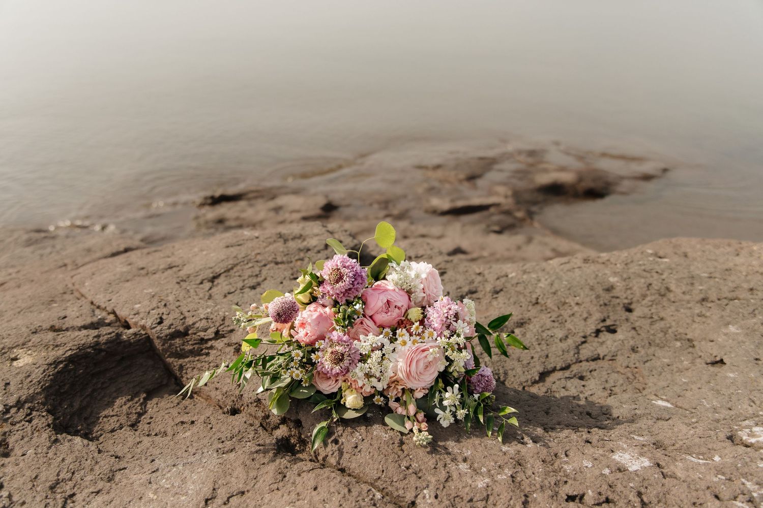 Pink rose bouquet resting on rocky shoreline with misty water in the background.