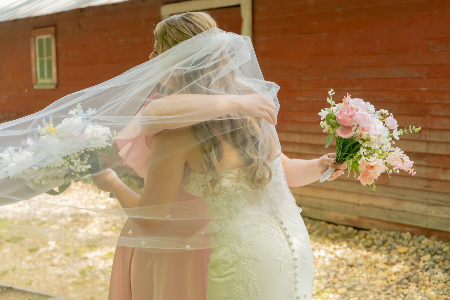 Bride in white gown and veil embracing bridesmaid in pink dress while holding floral bouquet near rustic red barn.