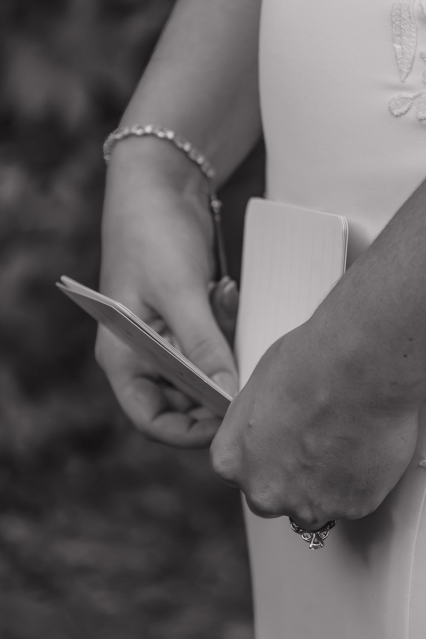 Black and white close-up of bride holding wedding vow booklet with pearl bracelet and ring visible.