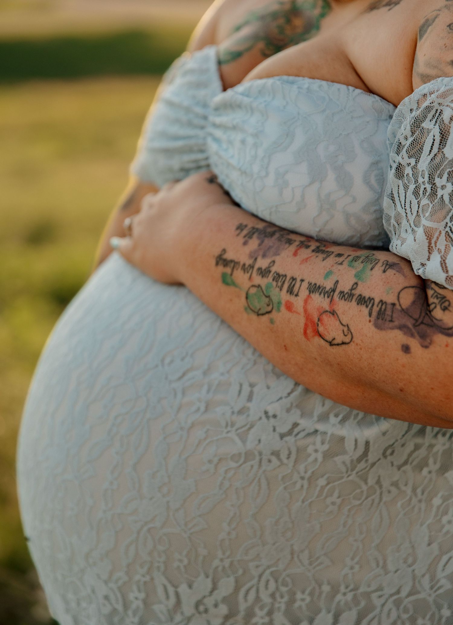 Pregnant woman in white lace dress with tattoos on her arm, cradling her baby bump outdoors in a field.