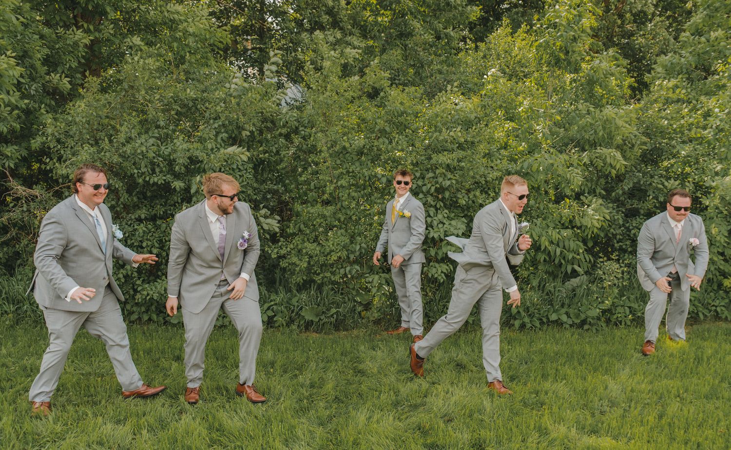 Five groomsmen in matching light gray suits and sunglasses strike fun poses on a green lawn at a wedding.