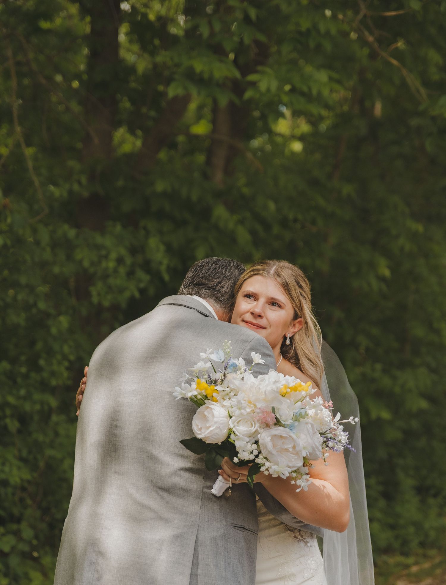 Bride in white gown and veil embracing a man while holding a white floral bouquet outdoors.