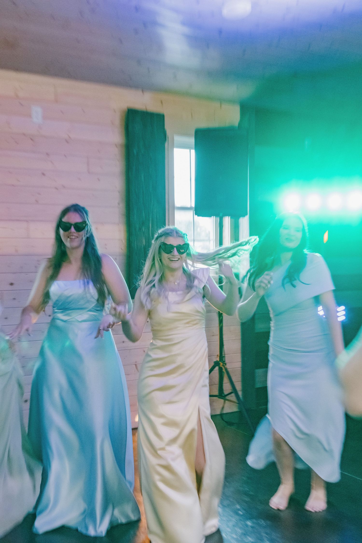 Bride and bridesmaids dancing under colorful lights at a lively wedding reception.