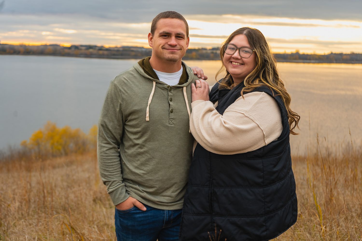 A couple poses together at sunset by a lake with autumn colors in the background.