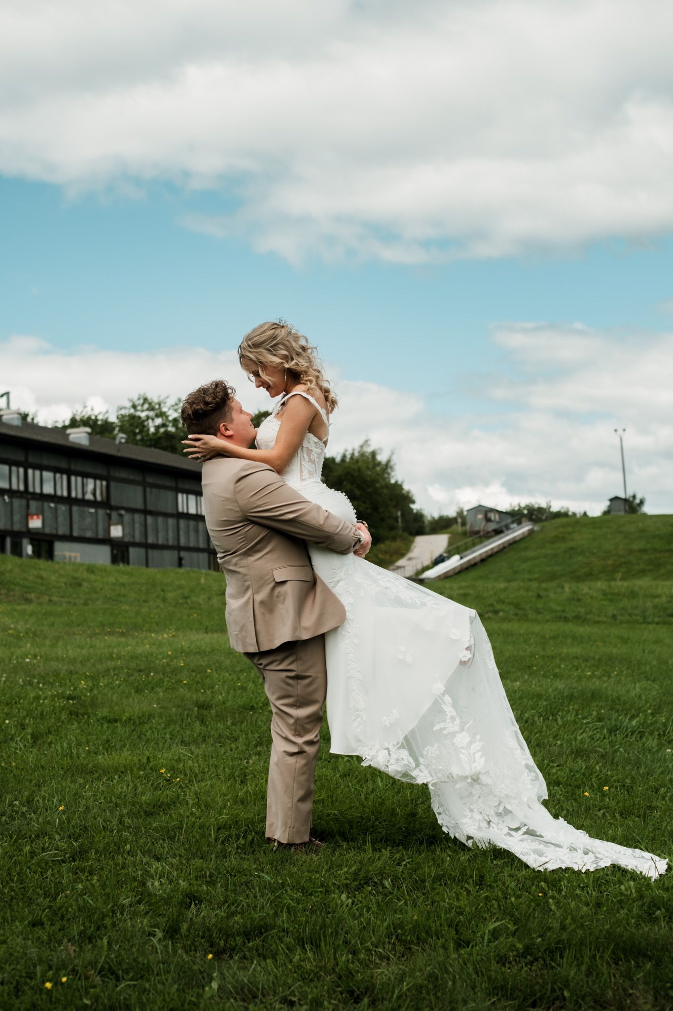Wedding couple shares playful moments on a grassy hillside with blue skies and clouds in the background.