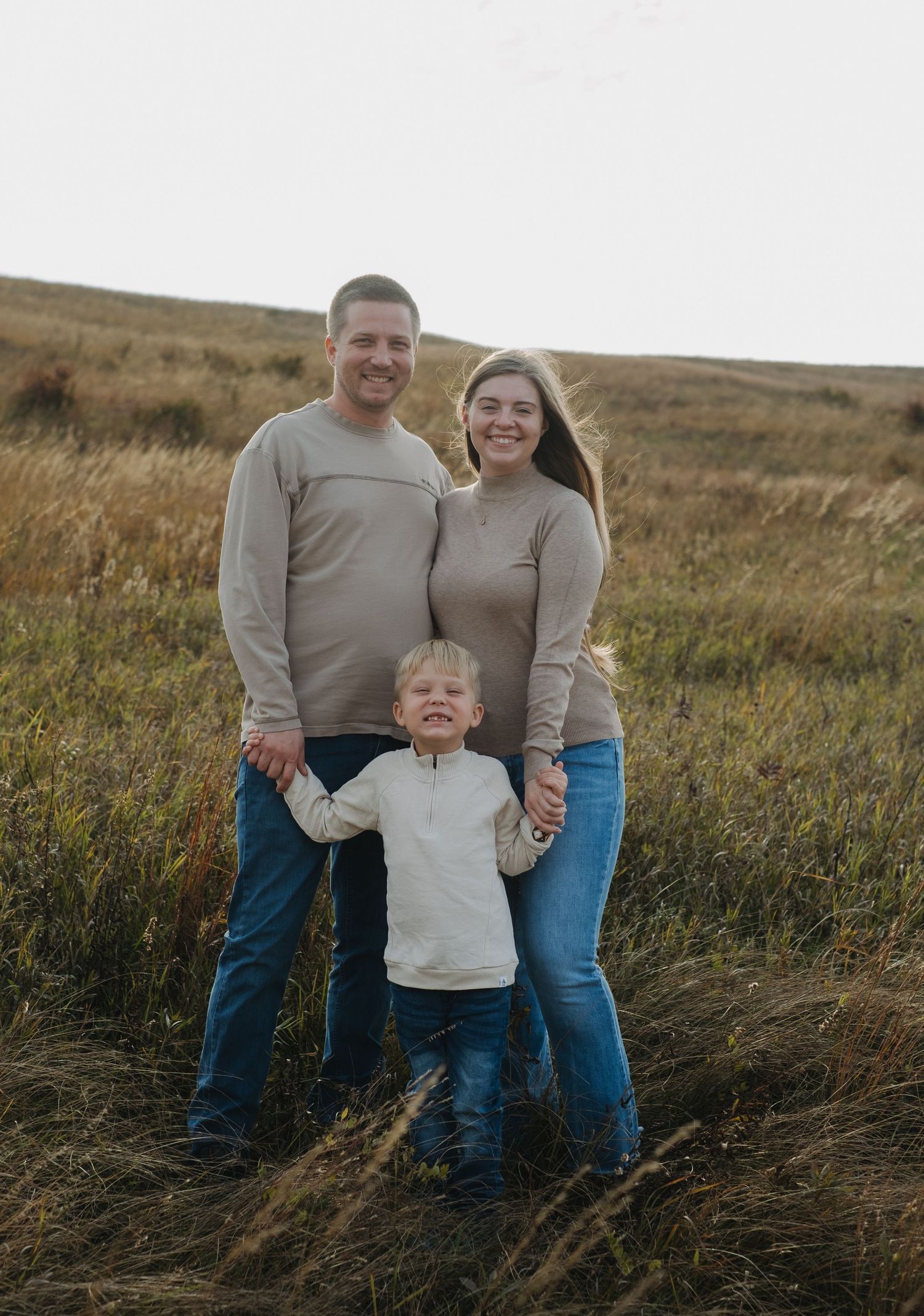 A family of three poses together in a grassy field at sunset.