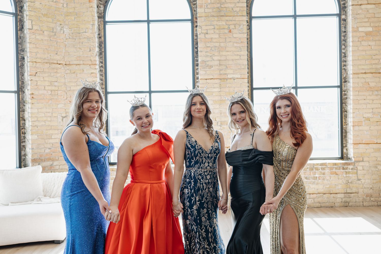 Five women in formal pageant gowns smile together in a bright loft space with brick walls.