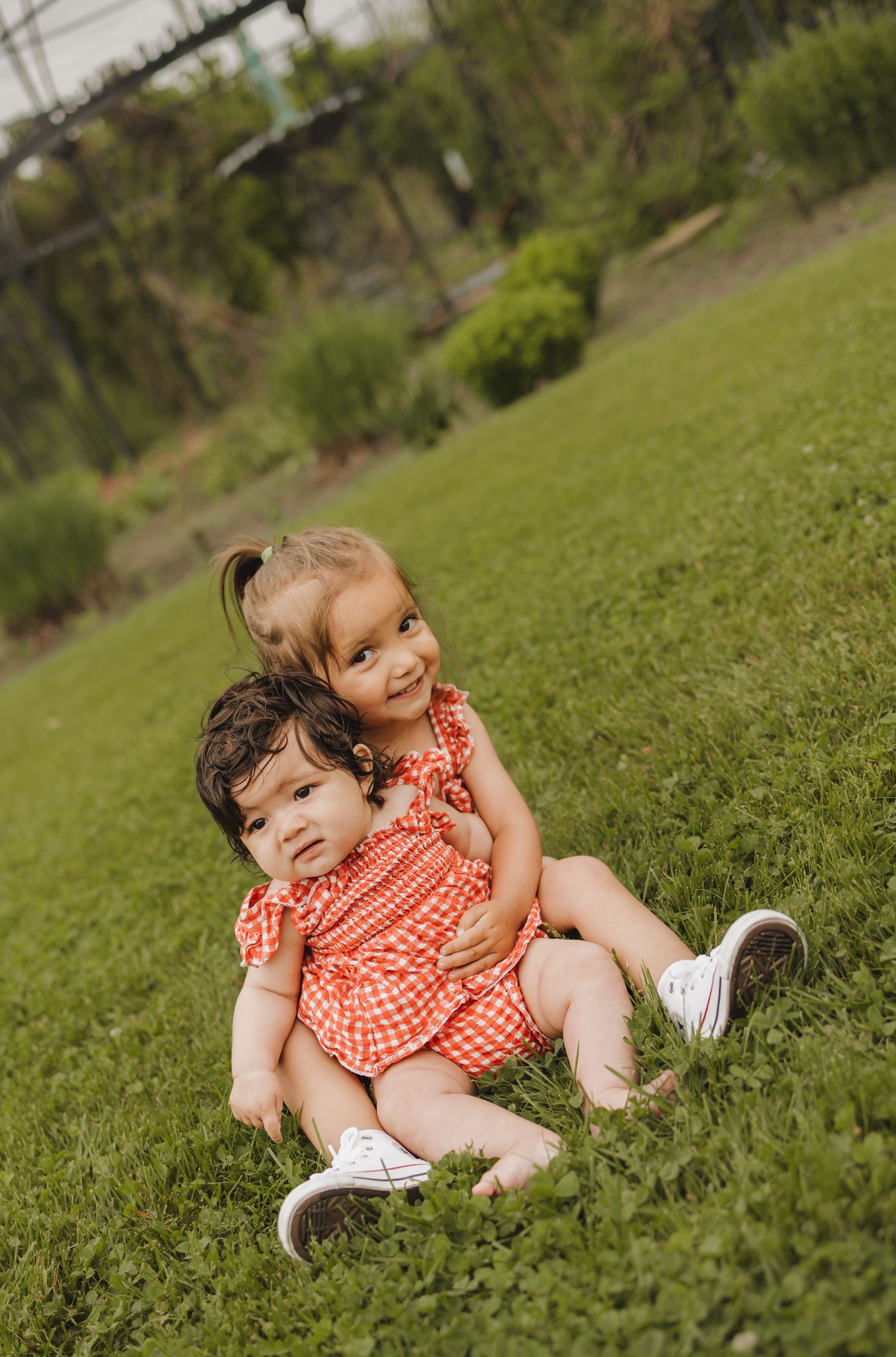 Two young sisters in matching red gingham outfits sit together on green grass in park.