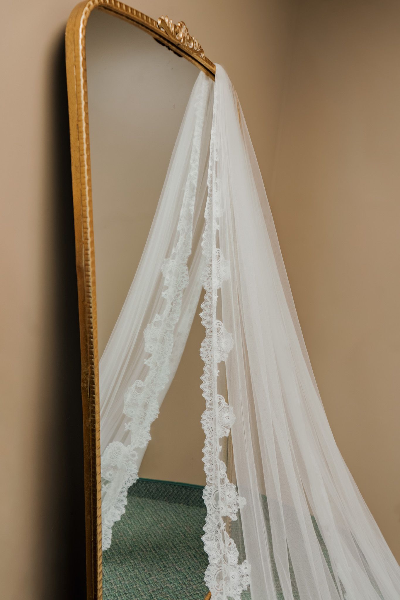 Elegant bridal veil with lace trim draped over a gold ornate floor mirror against a beige wall.