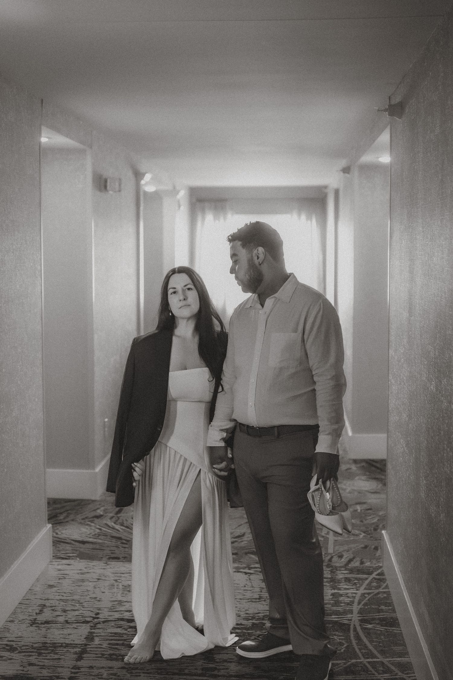 Black and white photo of a couple standing in a hotel hallway, woman in white dress with blazer, man in casual shirt.