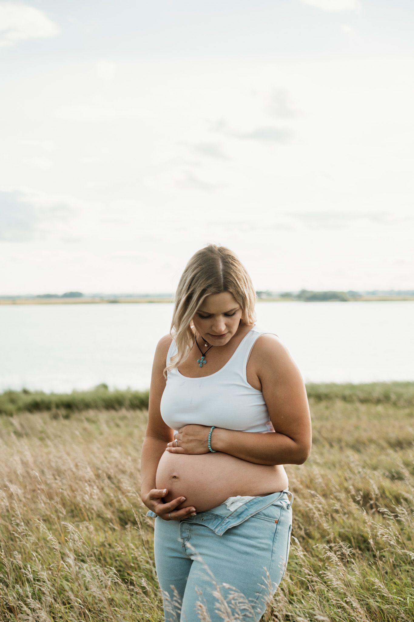 Pregnant woman in white tank top and jeans cradling her baby bump in a scenic field near water.