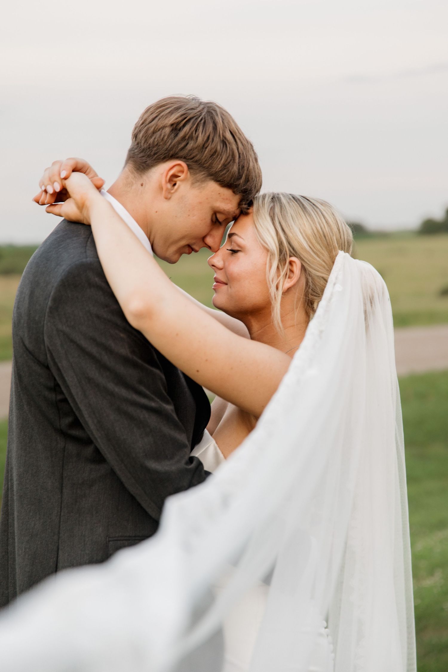 Bride and groom share an intimate forehead touch moment outdoors, with her veil flowing in the breeze.