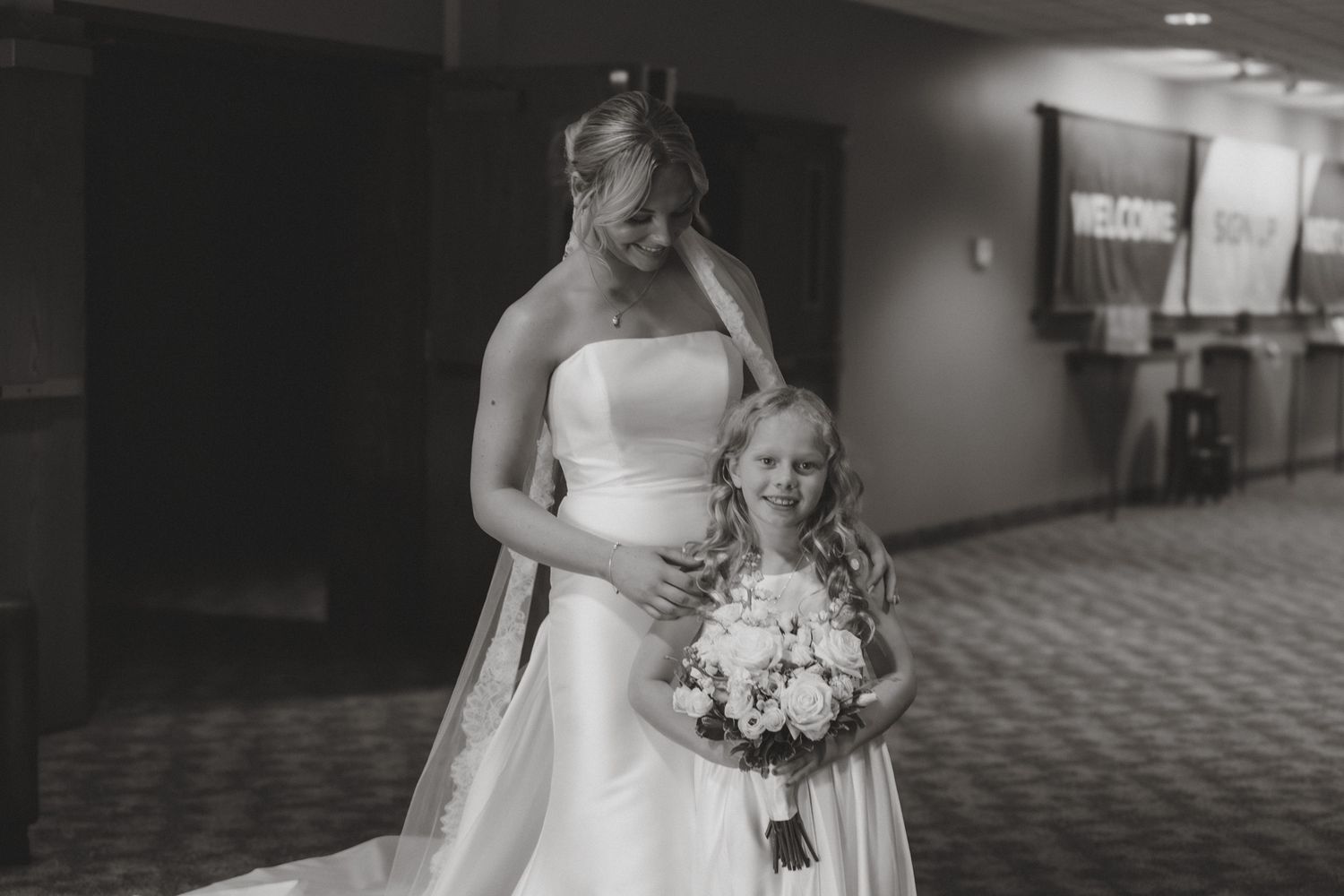 Black and white photo of a bride in a strapless gown posing with a young flower girl holding a bouquet.