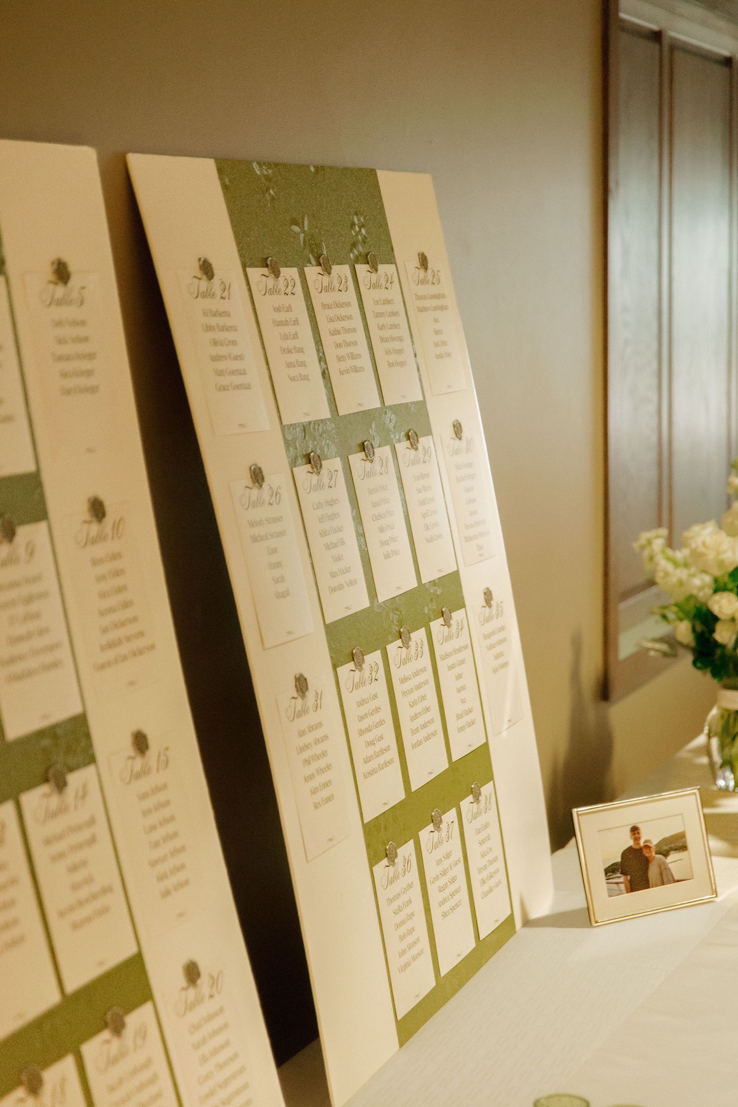 Wedding seating chart boards with green accents displayed on table beside floral arrangement and photo.