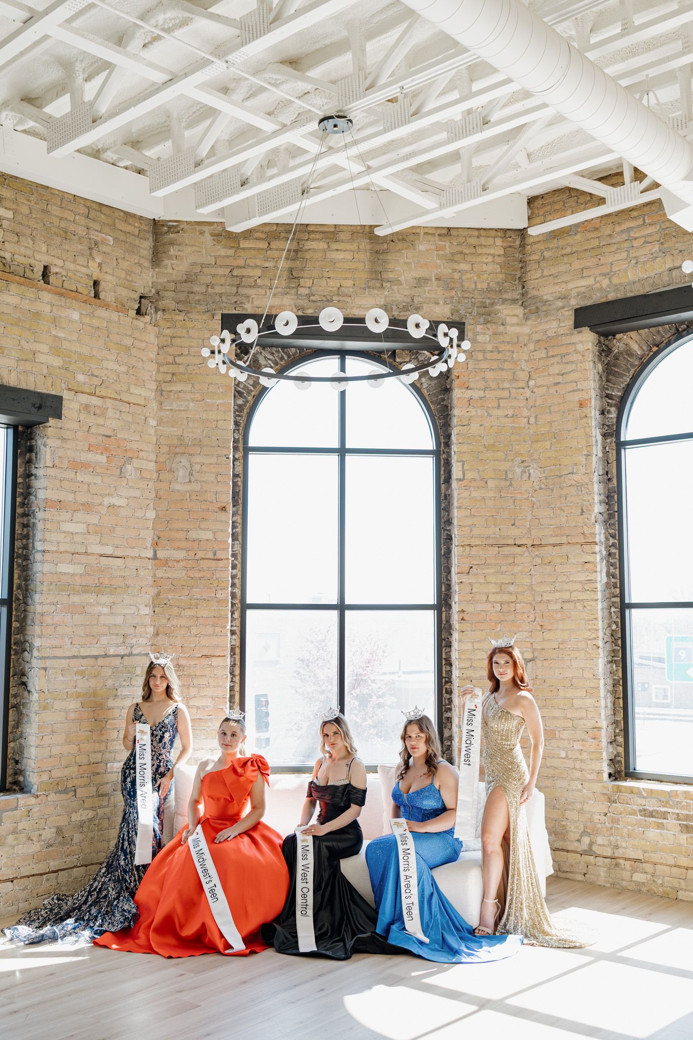 Five women in elegant pageant gowns and sashes pose in a rustic brick venue with arched windows and chandelier.