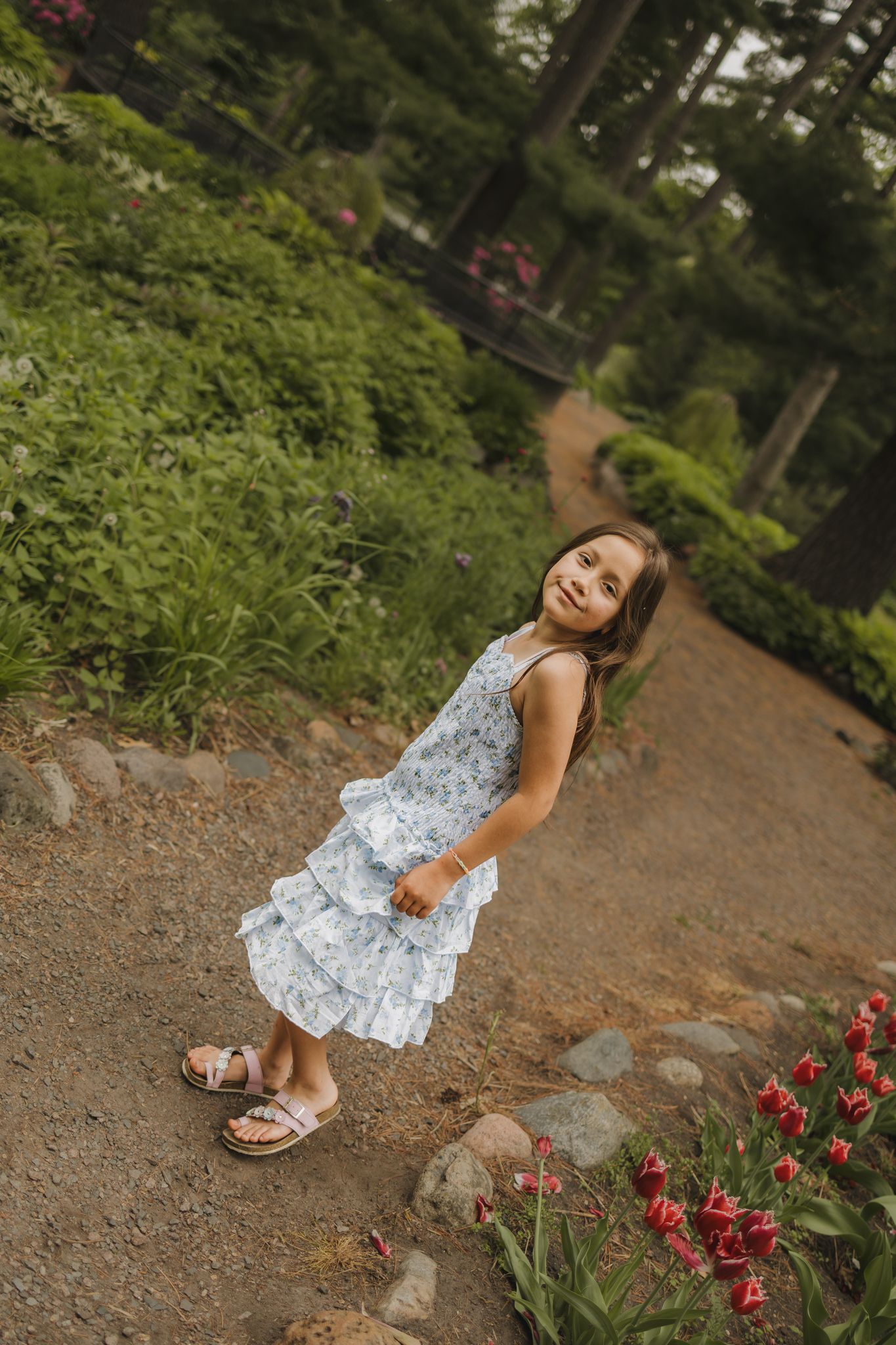 Young girl in floral white dress poses on garden path surrounded by lush greenery and red tulips.