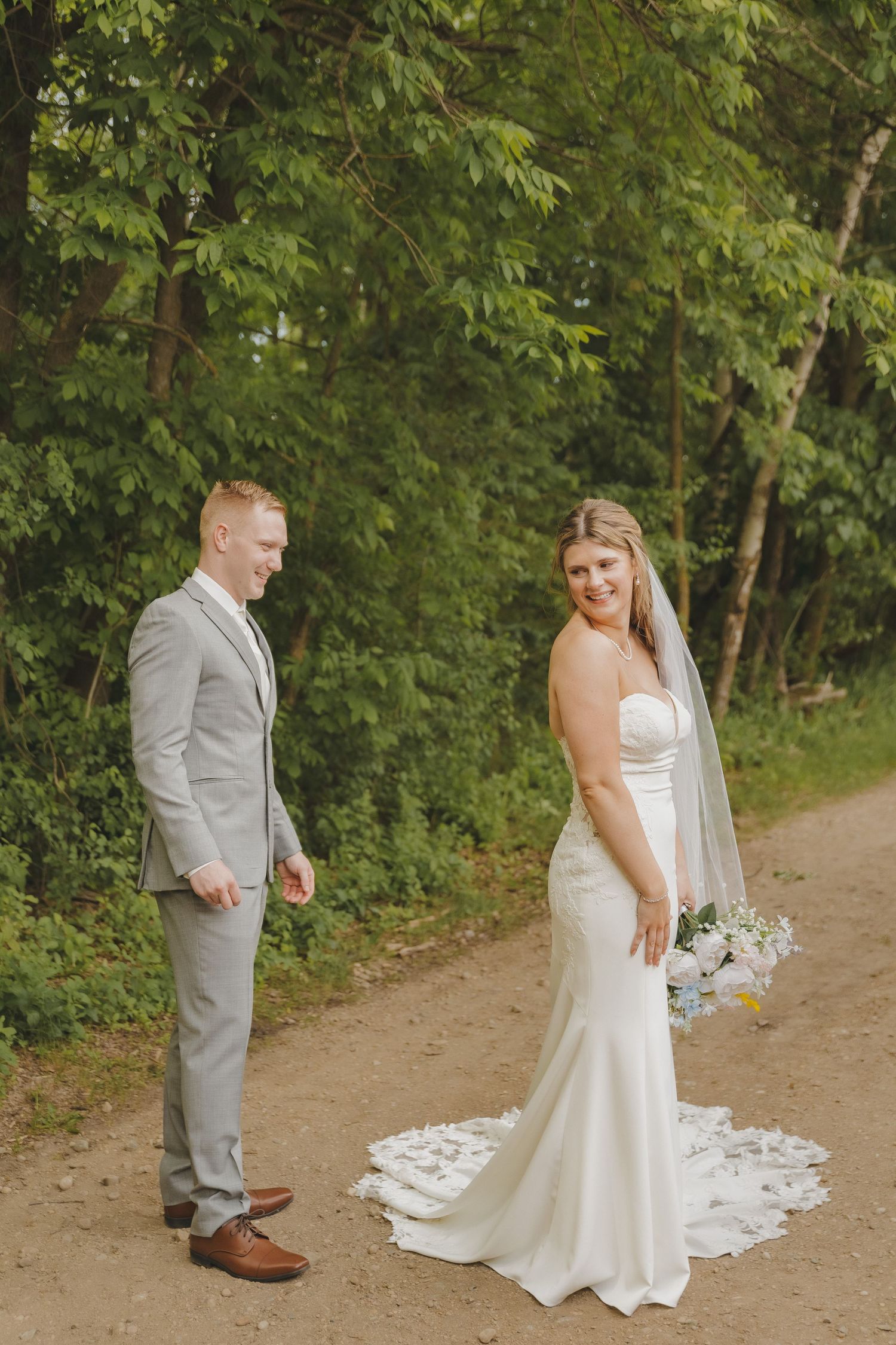 A bride in a white strapless gown and groom in a gray suit pose on a dirt path surrounded by lush green trees.