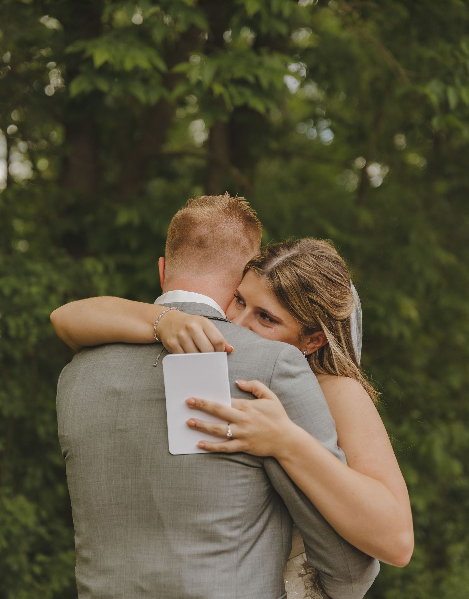 Emotional bride in white gown embraces groom in gray suit outdoors during heartfelt first look moment.