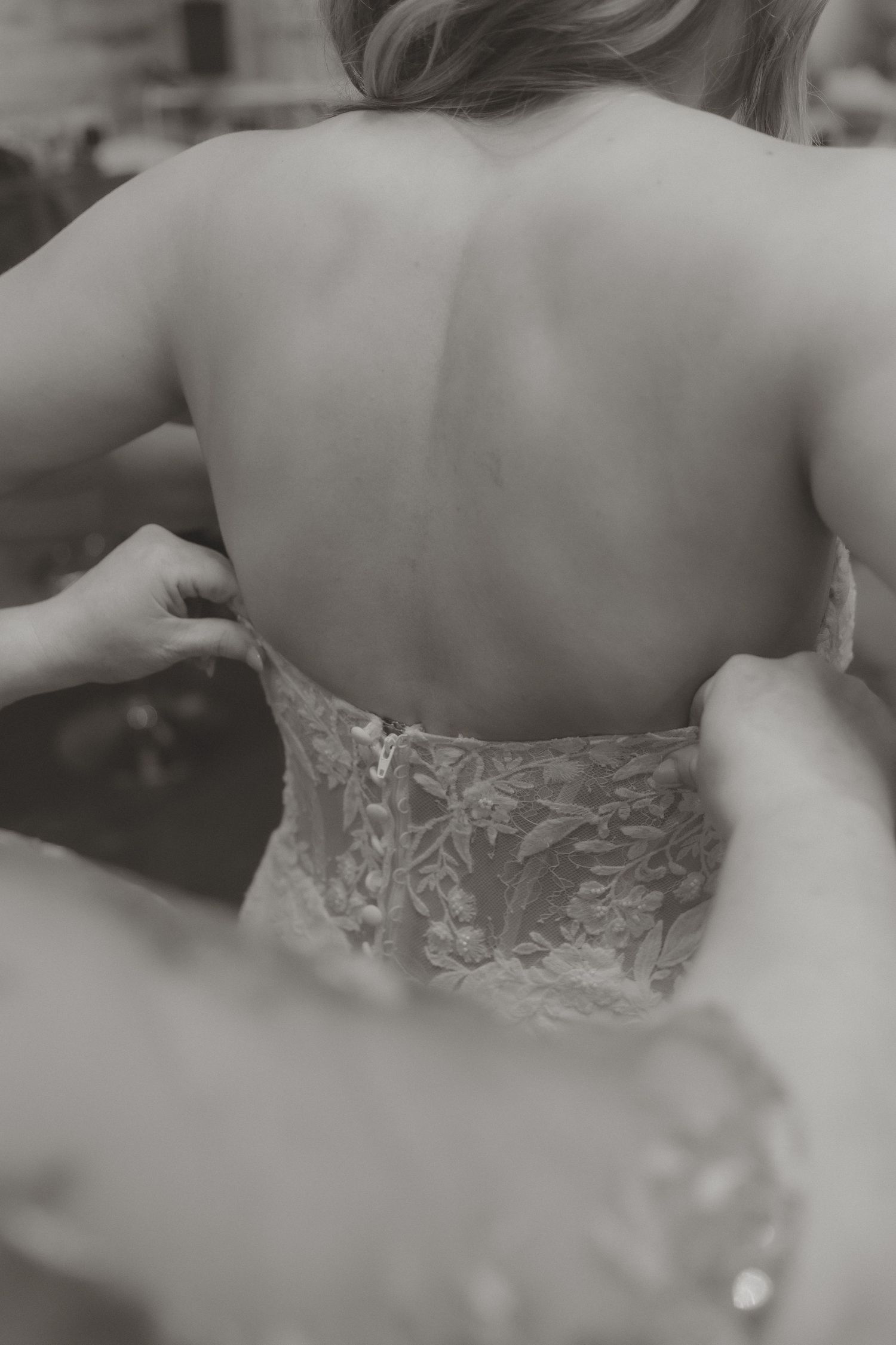 Black and white photo of a bride having her lace wedding dress fastened from behind, showcasing intricate floral detailing.