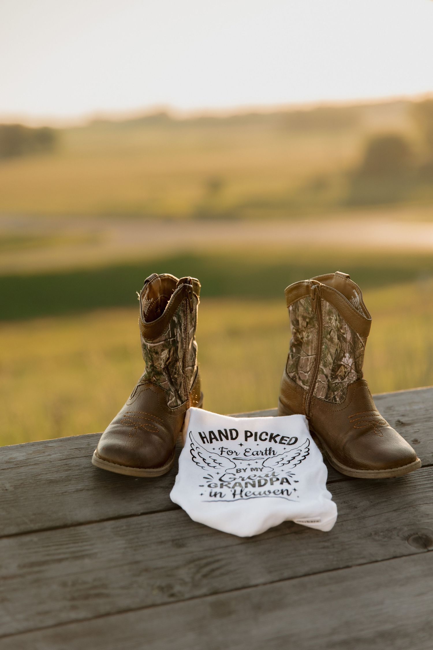 Cowboy boots with camo tops beside a onesie reading Hand Picked for Earth by my Grandpa in Heaven.