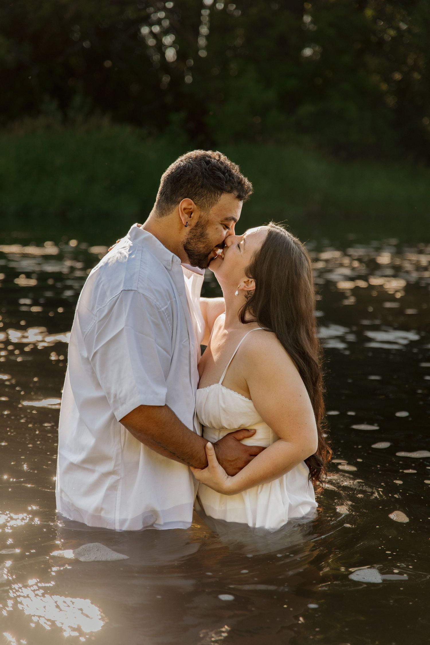 A couple in white clothing shares a romantic kiss while standing in a sunlit river surrounded by lush greenery.