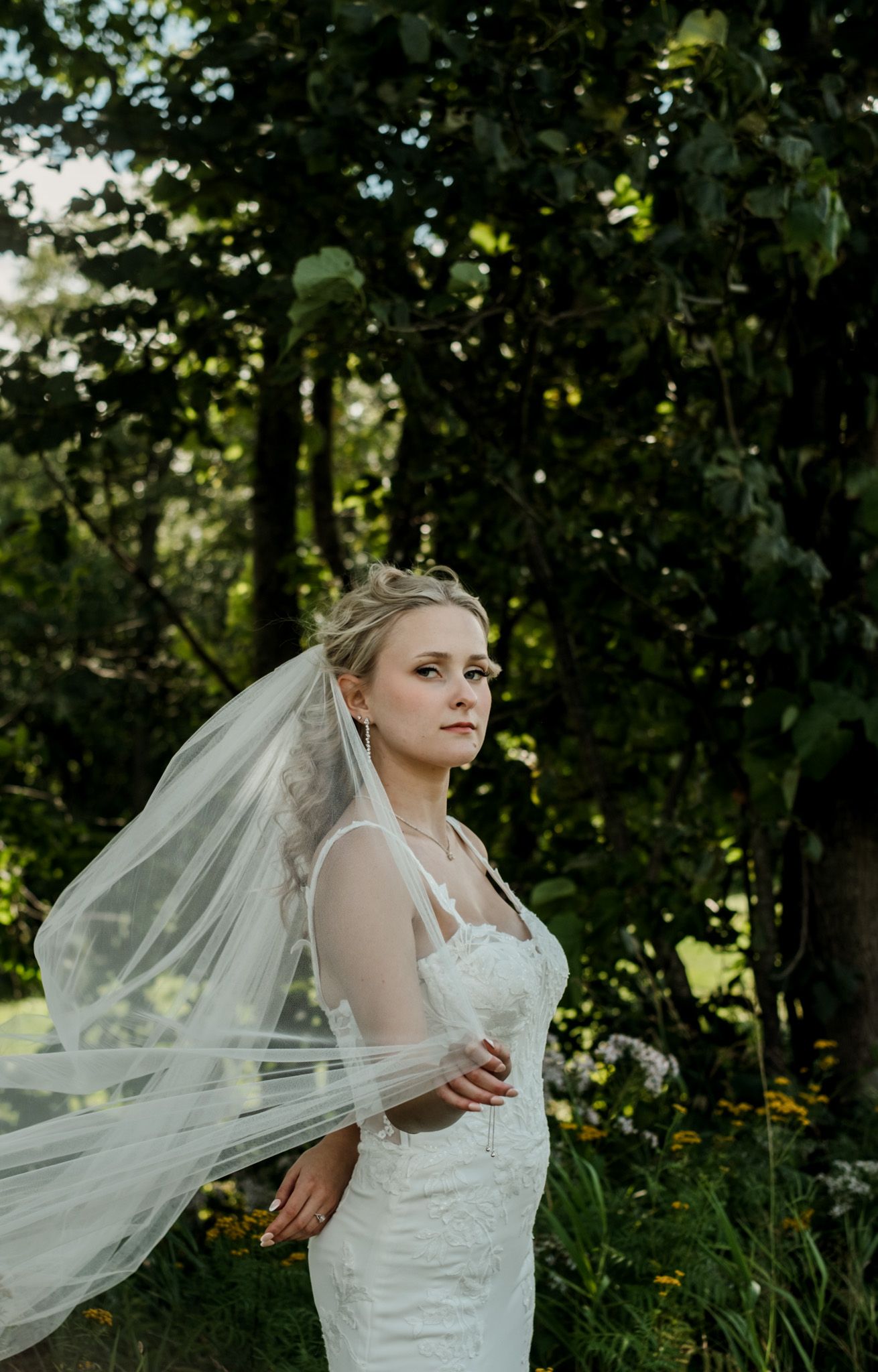 A bride in a flowing veil poses gracefully against a backdrop of lush green foliage.