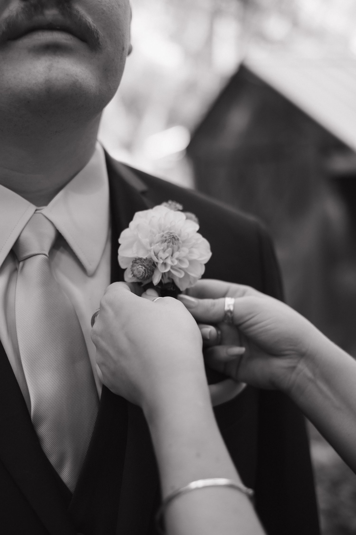 Hands pinning white floral boutonnieres onto dark suit lapels in black and white.