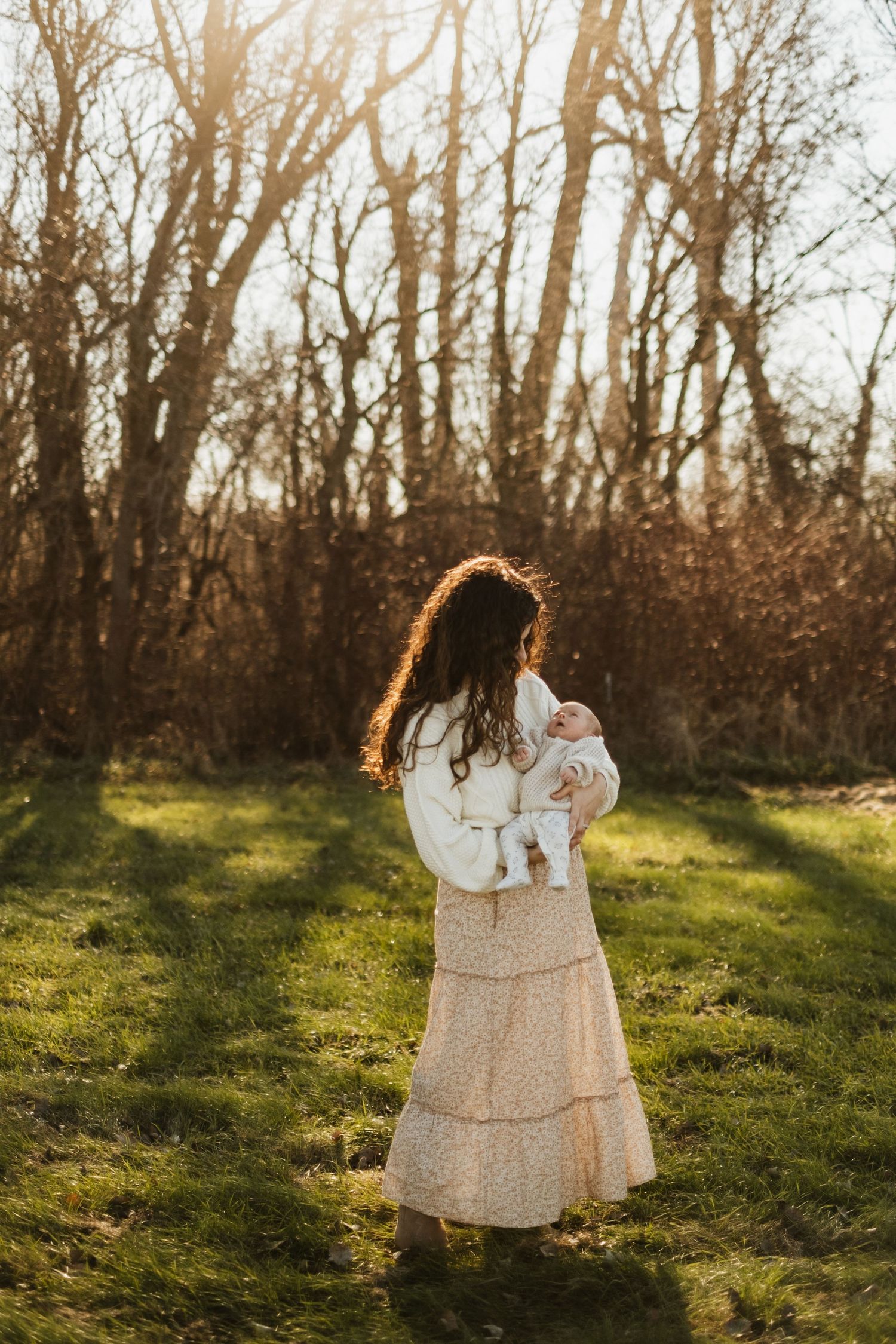 A woman in a white sweater and long skirt holds a baby while standing in a sunlit forest clearing.