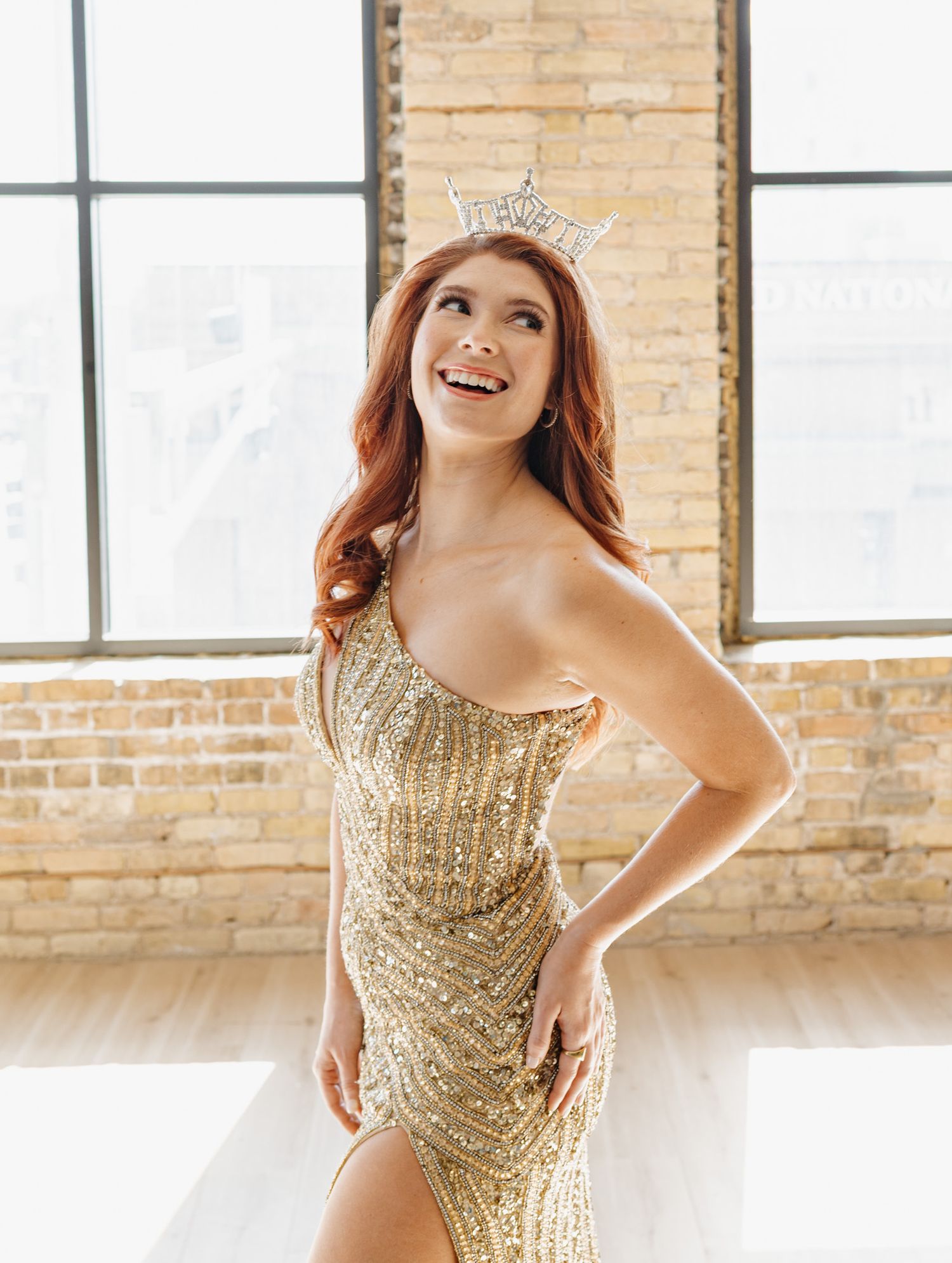 Woman in gold sequin gown and rhinestone crown smiling in brick loft venue.