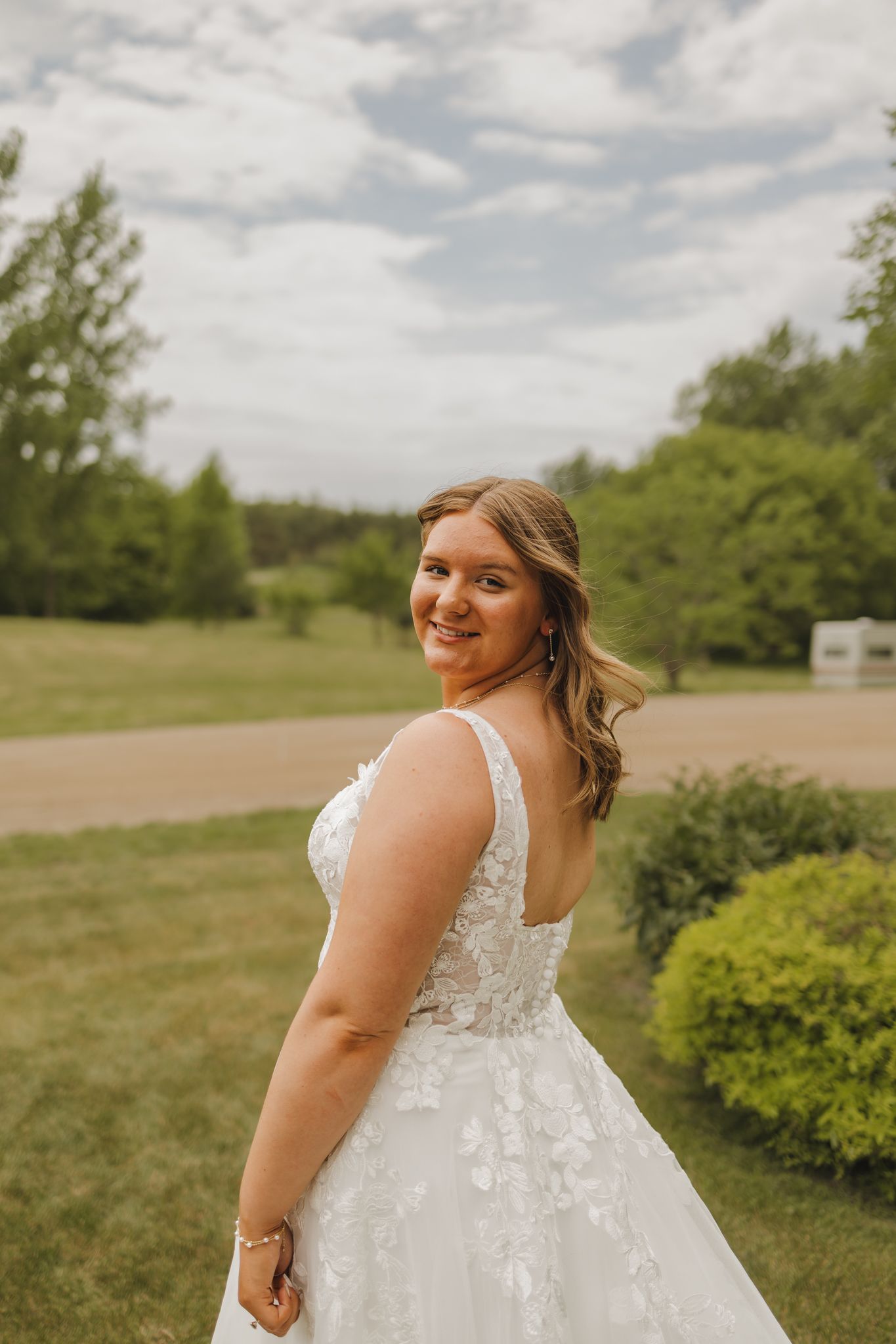 Bride in a floral lace wedding gown smiling over her shoulder in an outdoor garden setting.