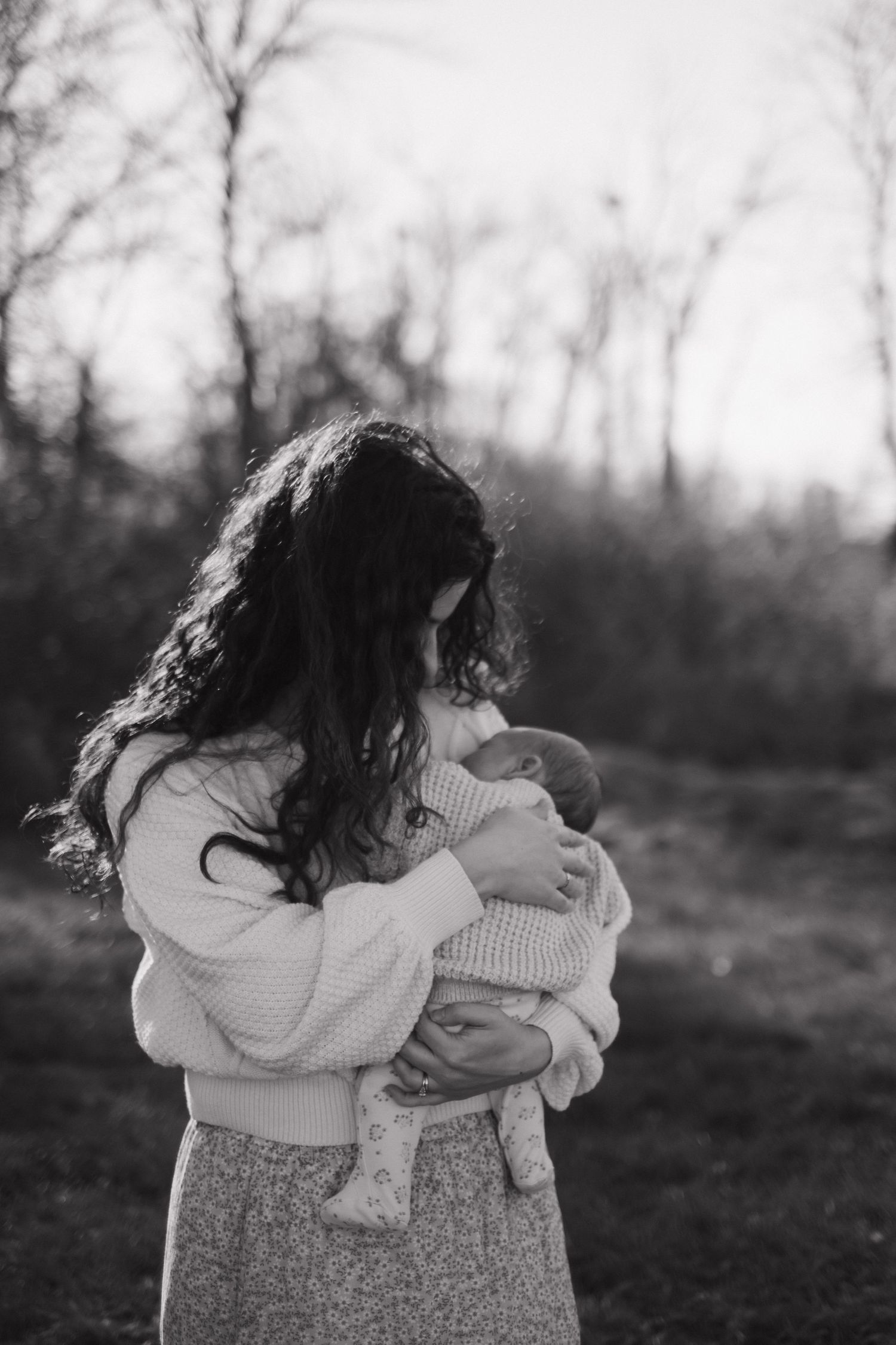 Black and white photo of a mother cradling her newborn baby outdoors among bare winter trees.