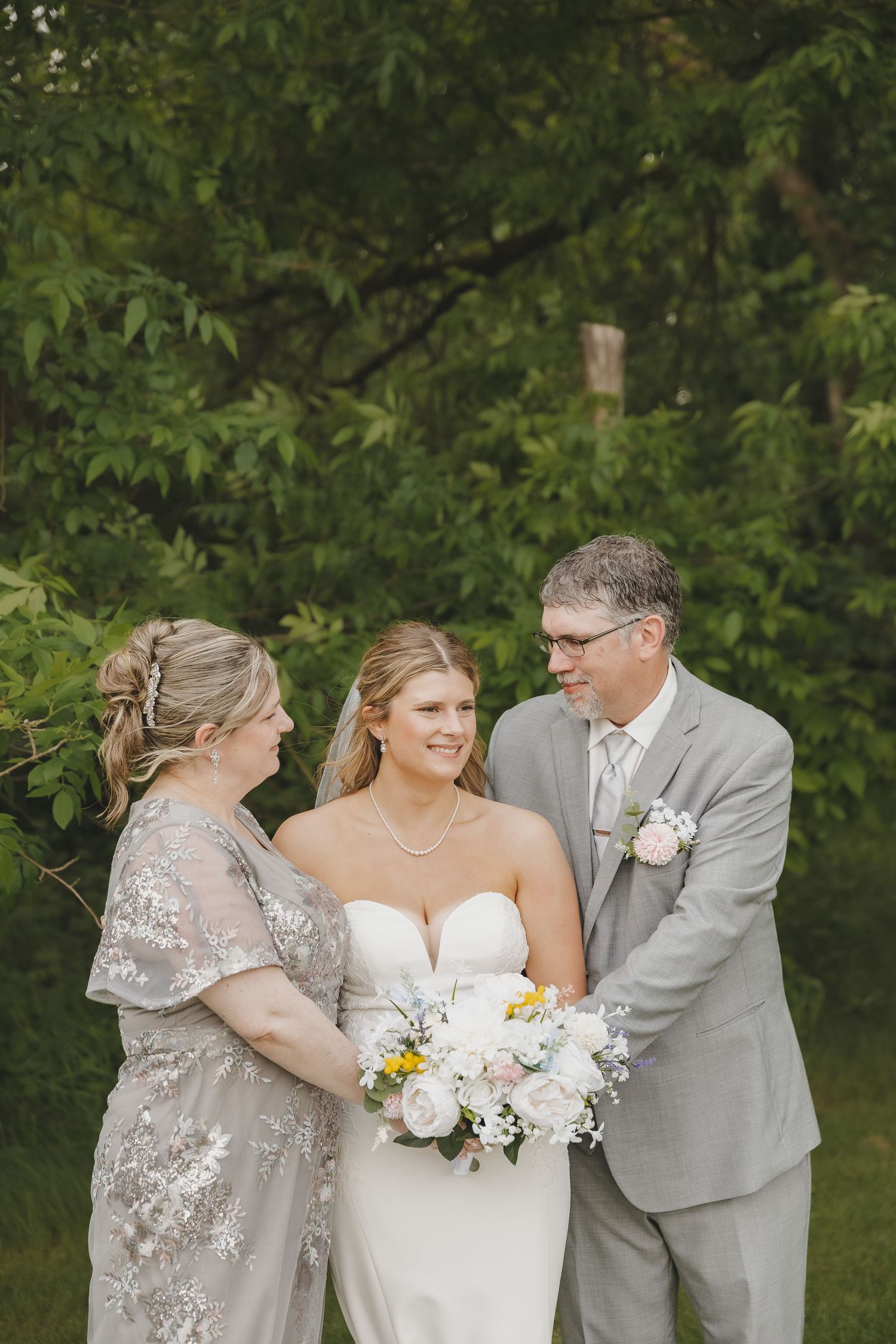 Bride in strapless gown holds bouquet flanked by parents in gray attire outdoors.