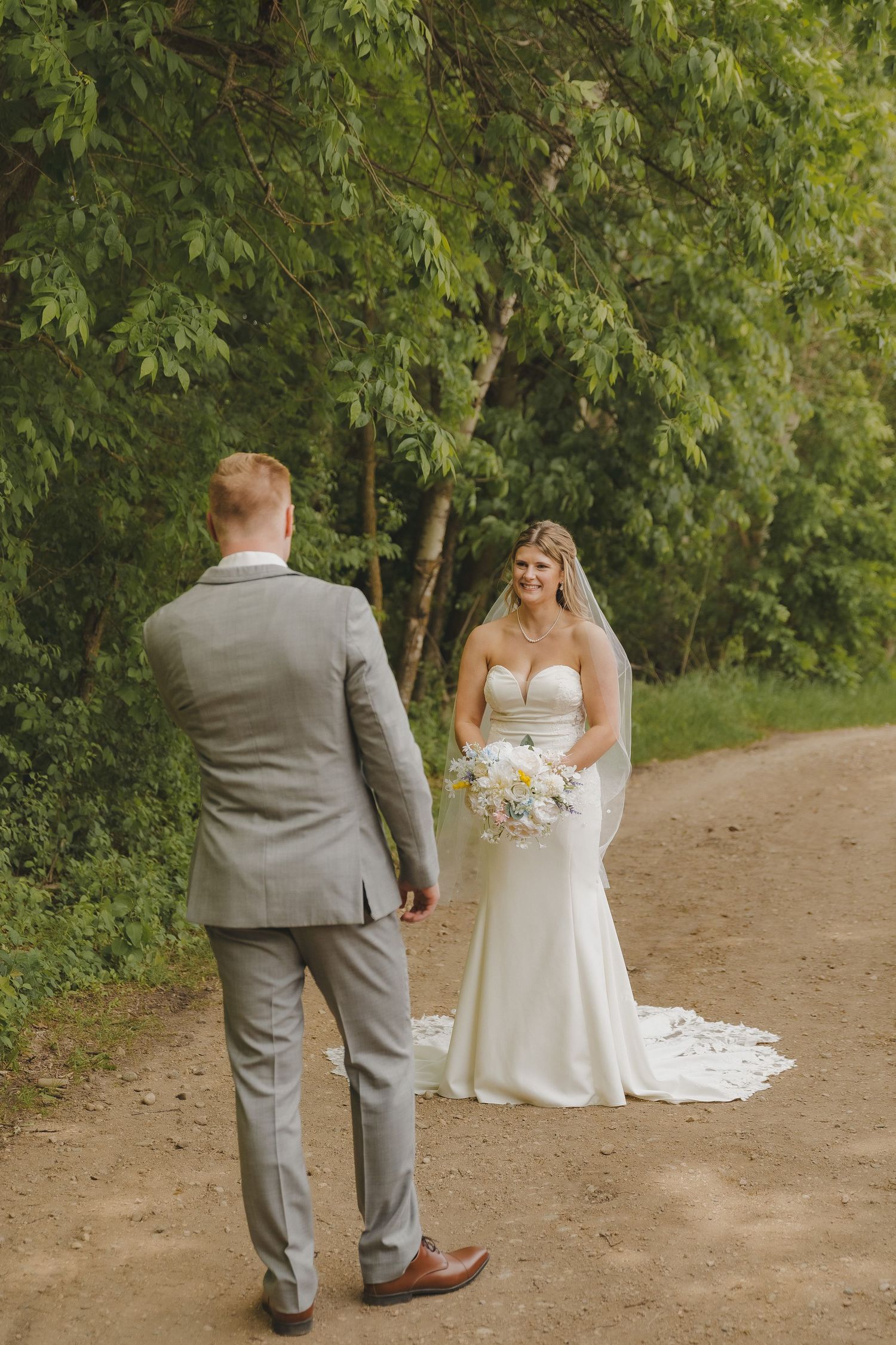 Bride in white gown holding bouquet smiles at groom on a tree-lined dirt path.