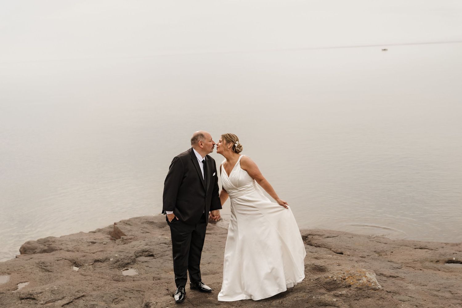 Bride and groom sharing a romantic kiss on rocky shoreline with misty lake backdrop on their wedding day.