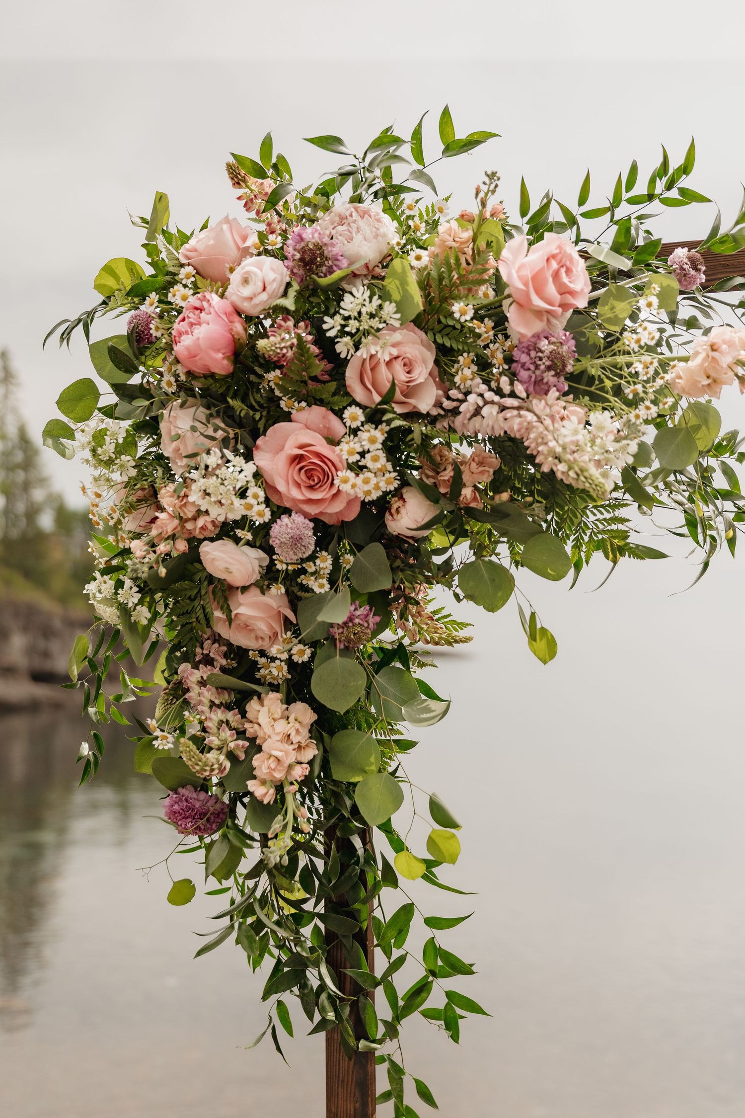 Elegant wedding arch floral arrangement with pink roses, greenery, and wildflowers against a misty waterfront.