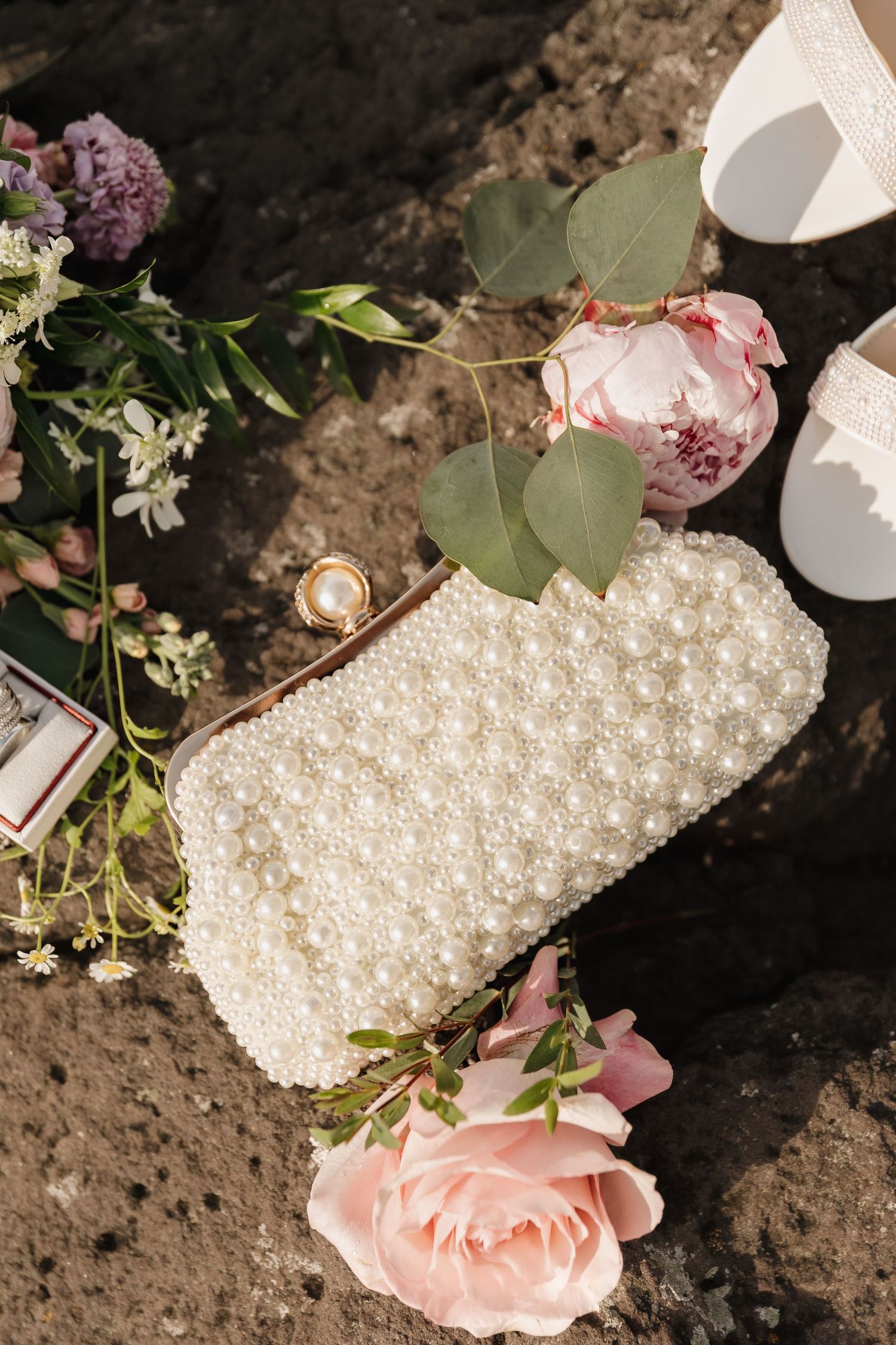 Pearl beaded bridal clutch purse surrounded by pink roses, eucalyptus, gold rings, and white wedding heels on stone surface.