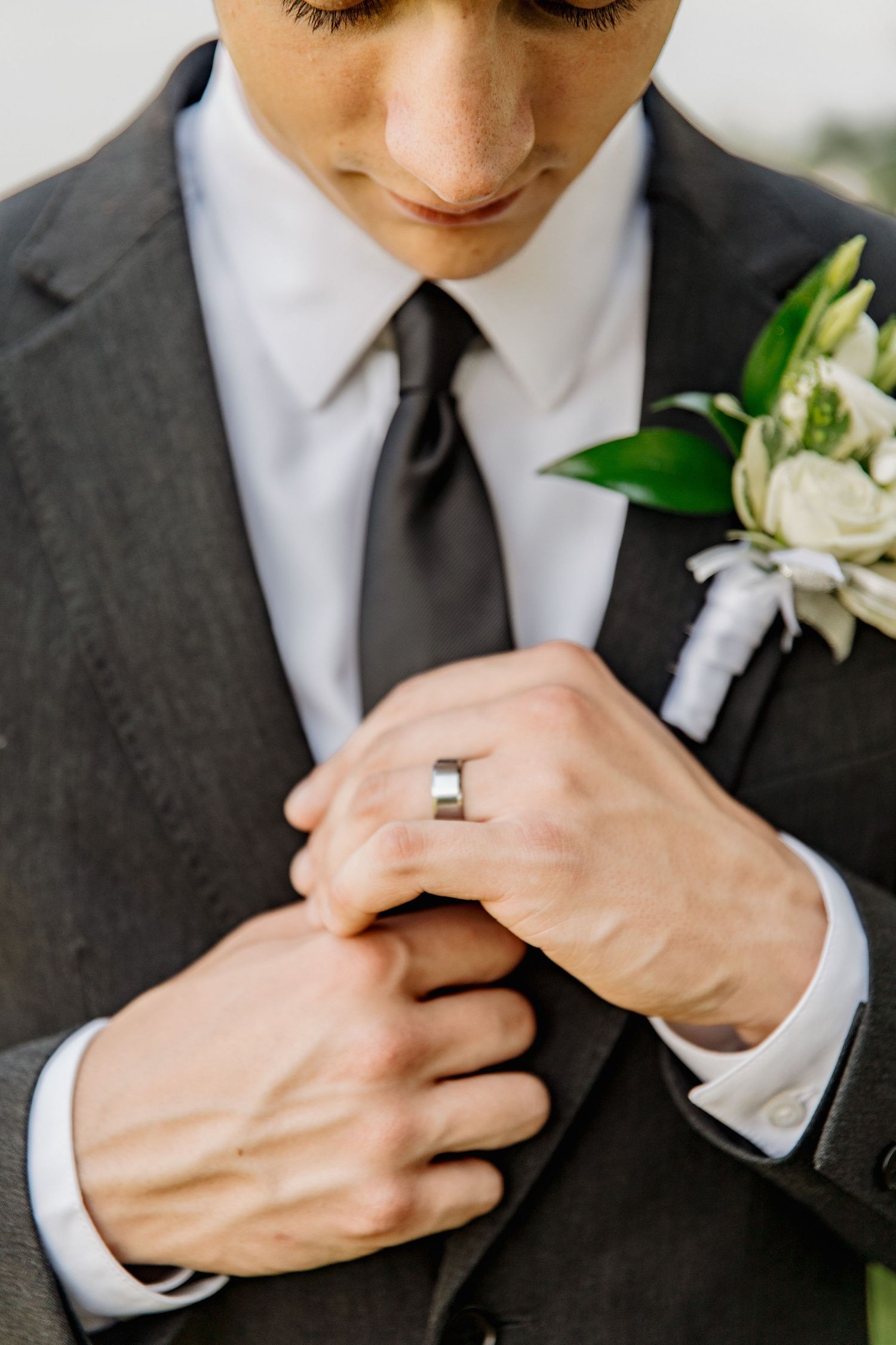 Groom in gray suit adjusting dark tie with wedding ring visible, white floral boutonniere on lapel.