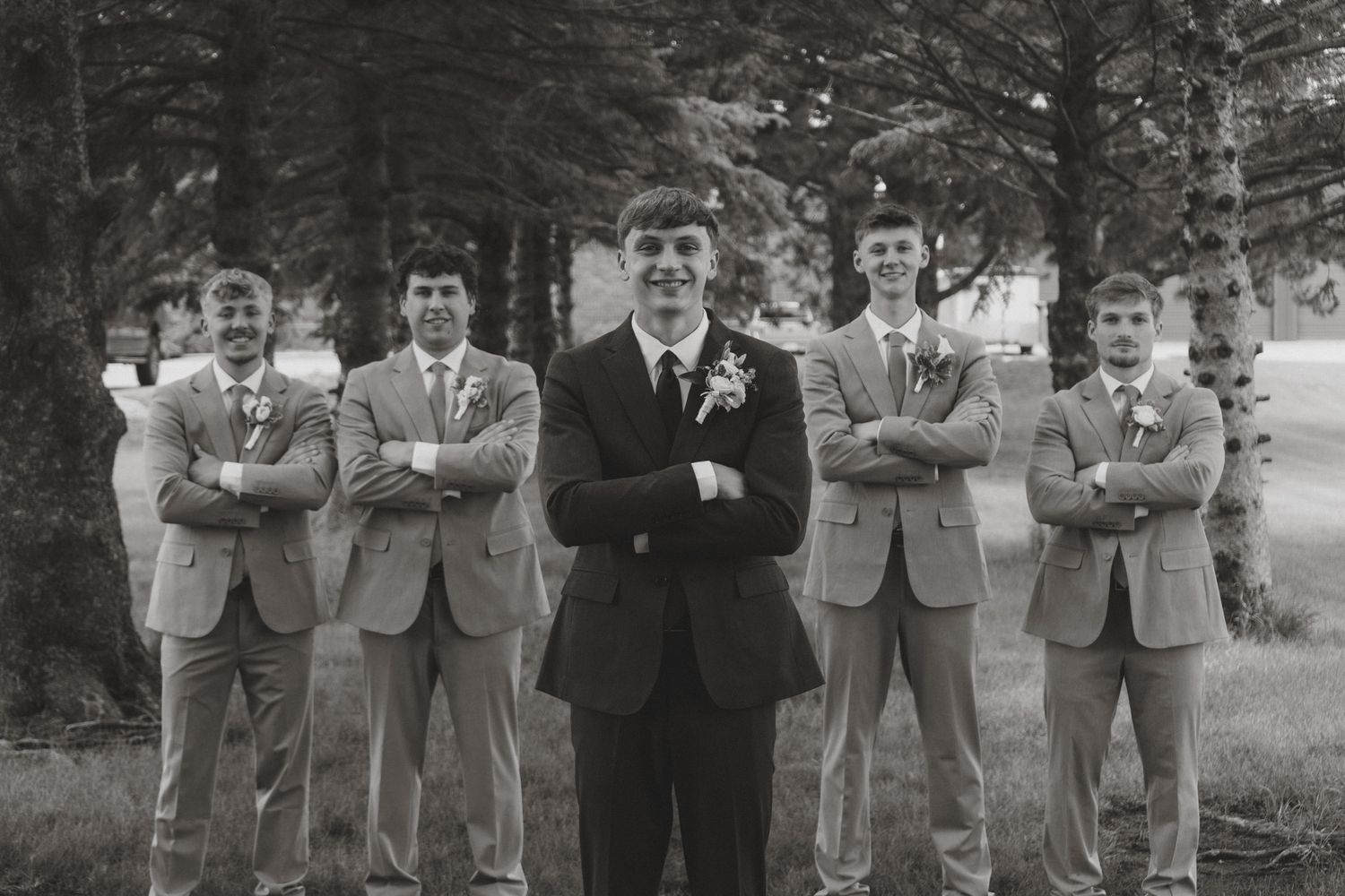 Black and white photo of groom with groomsmen in suits, arms crossed, posing outdoors at a wedding.