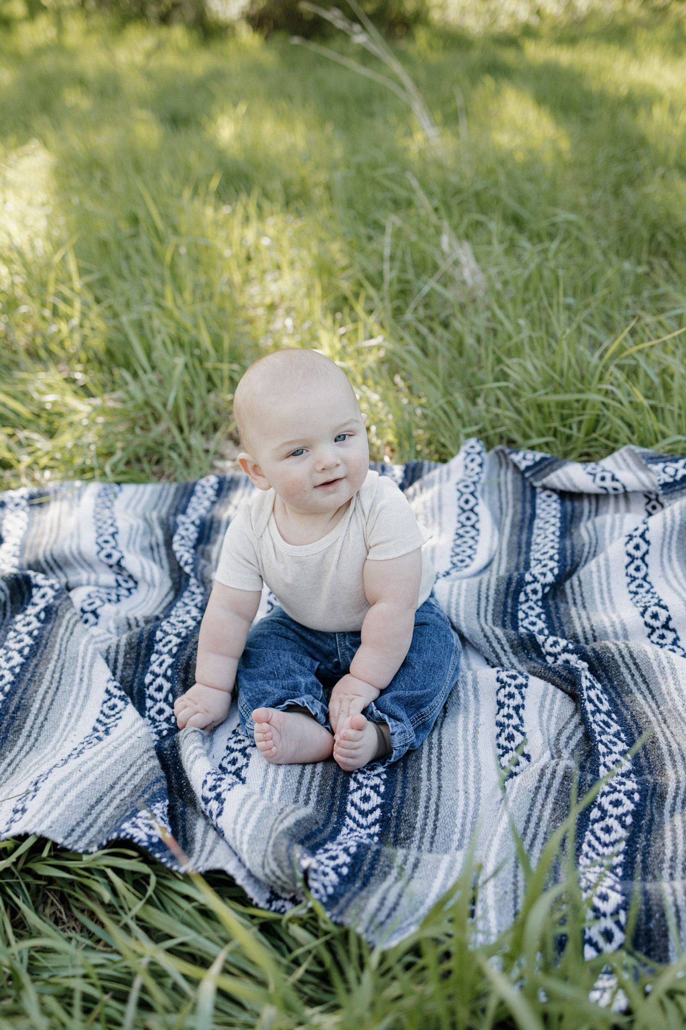 Baby in white onesie and jeans sitting on blue patterned blanket in green grass outdoors.