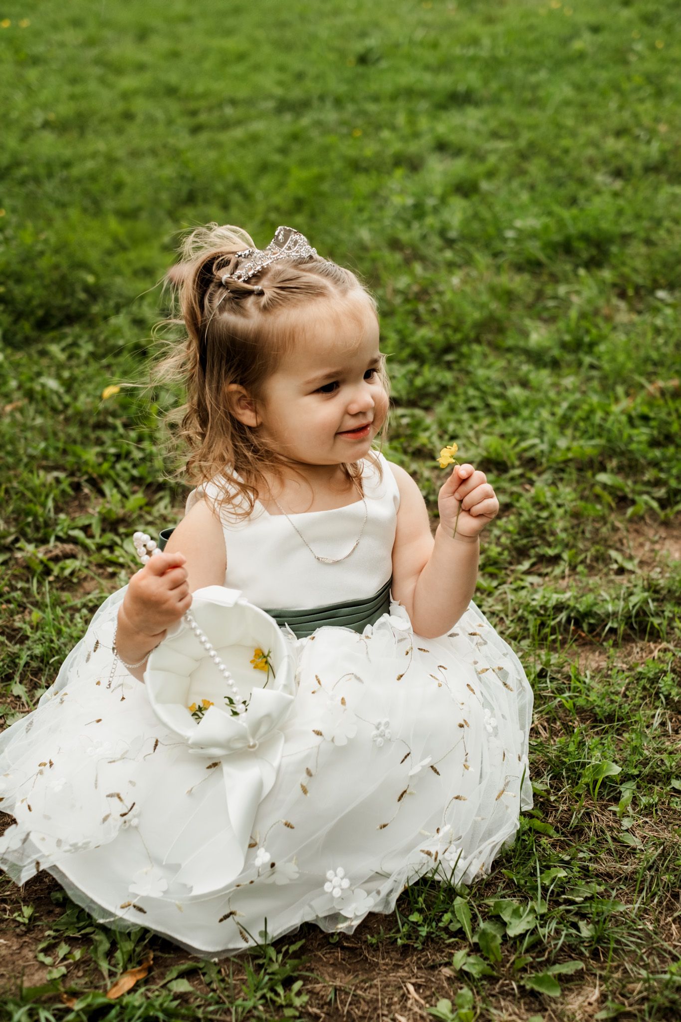 Flower girl in white dress plays joyfully on green grass during wedding.