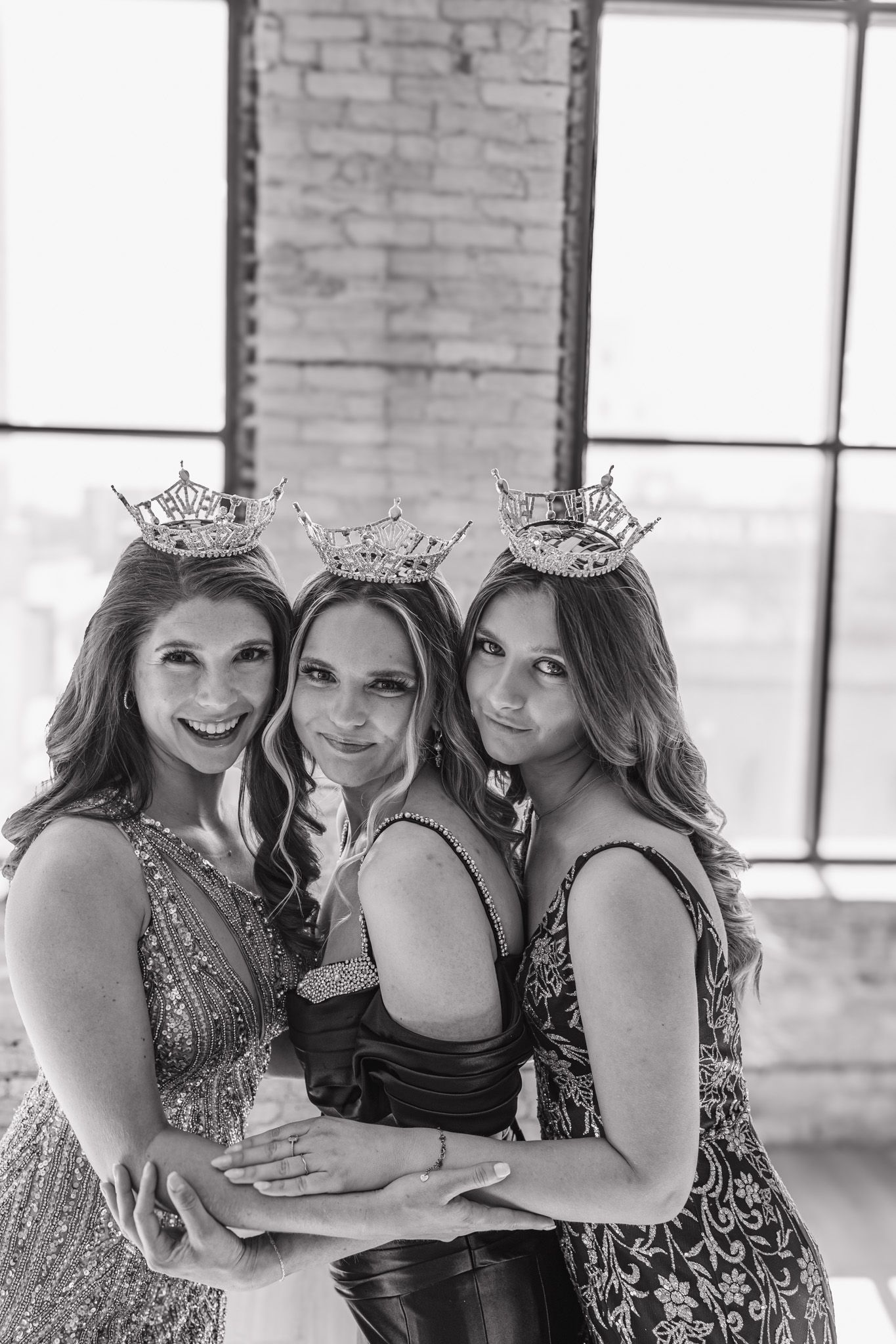 Three pageant queens wearing sparkling crowns embrace in a black and white portrait.